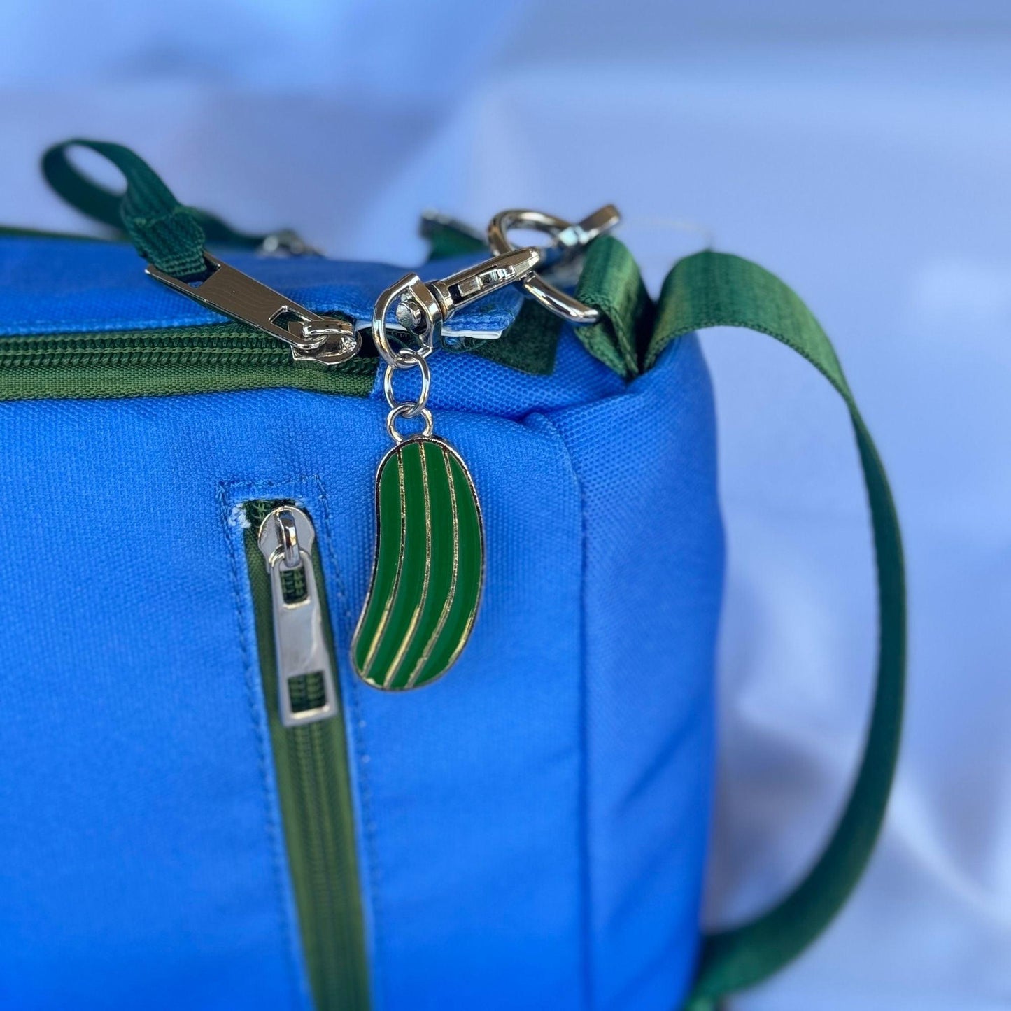 A close-up of the Fresh Pickle Insulated Cooler Bag in blue, featuring green zippers and straps. A metallic keychain shaped like a green pickle with gold edges hangs from the zipper. Background is softly blurred in light blue and white.