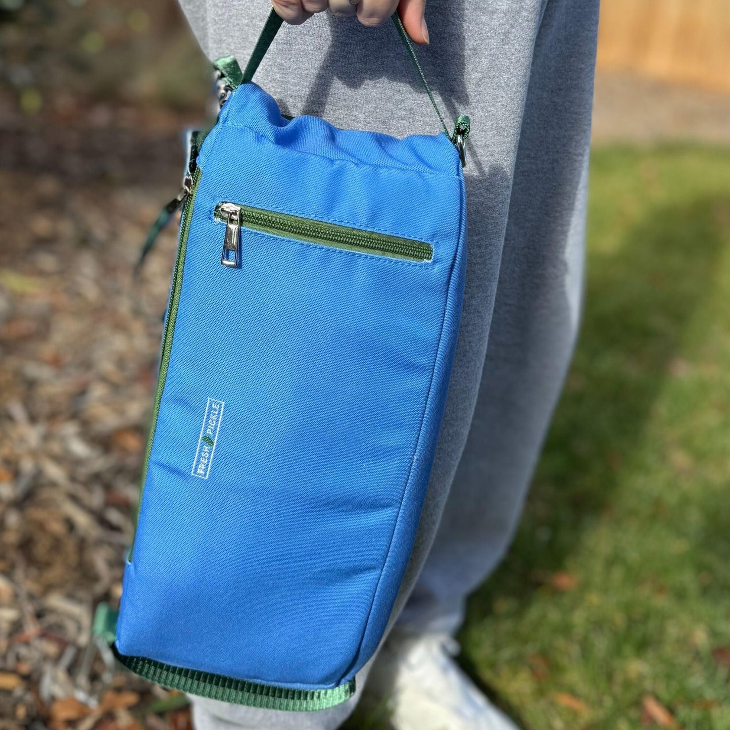 A person in gray sweatpants and white shoes holds the Fresh Pickle Insulated Cooler Bag by its green handle. The bright blue bag has a front zipper pocket, green trim, and is perfect for court essentials. Grass and mulch appear in the background.