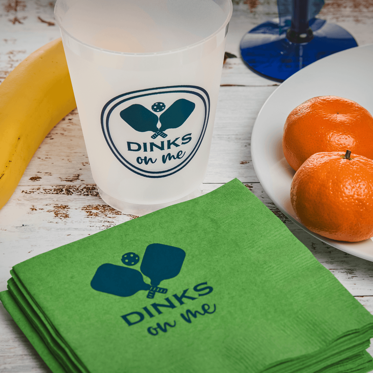 A Fresh Pickle Pickleball Cocktail Napkin and a white cup, both reading “DINKS on me” with crossed paddles, sit on a wooden table next to a banana, two clementines on a plate, and the base of a blue wine glass.