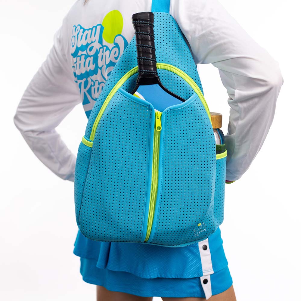 A person is seen from behind with the Swinton Pickleball Sling Bag in blue, featuring neon yellow accents. They hold a tennis racket and water bottle, wearing a blue skirt and a white long-sleeve shirt with "Stay Outta the Kitchen" on the back.