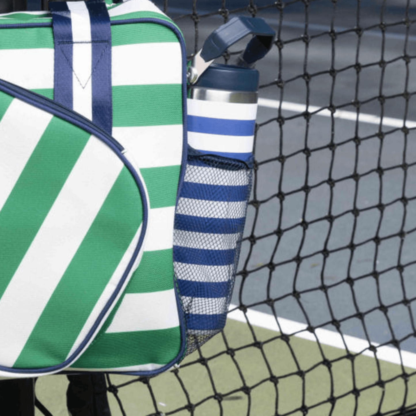A green and white striped bag with navy accents hangs on a tennis net, featuring a rounded zipper pocket. In the side mesh pocket is the Fresh Pickle Tumbler by Fresh Pickle, a 30 oz water bottle with a flip-up straw. Blue and green tennis court in background.