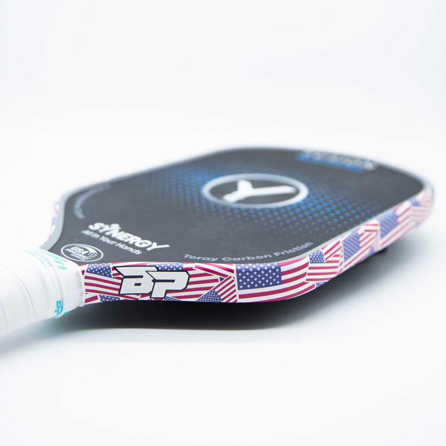 A close-up of a black pickleball paddle with a white handle features Bodhi Performance's premium edge tape decorated with small American flags, providing professional edge guard protection. Branding and text appear on the paddle.
