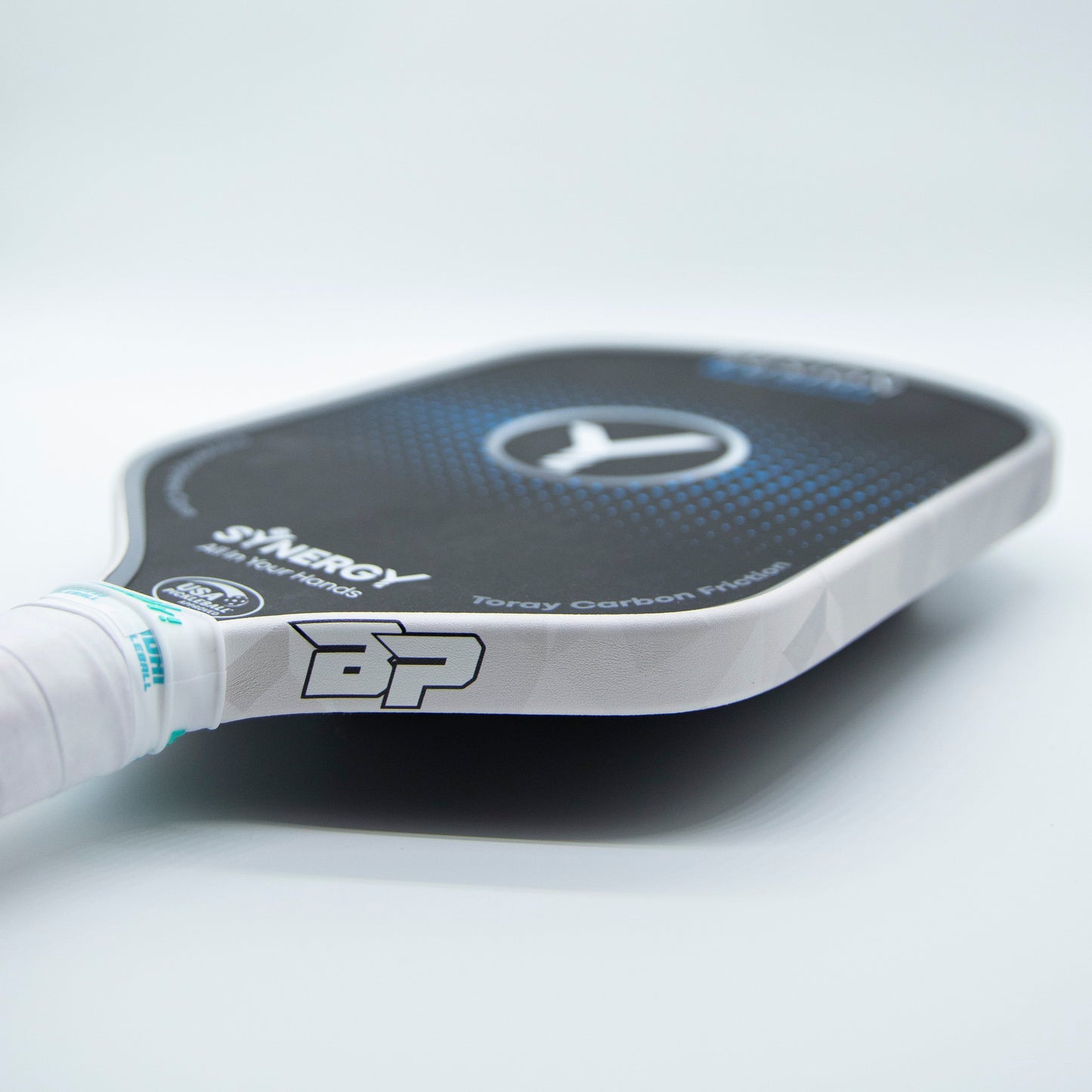 Close-up of a black and white pickleball paddle with a textured face, SYNERGY branding, BP logo, and handle wrapped in white grip tape. Bodhi Performance Premium Edge Tape provides professional edge guard protection against wear.