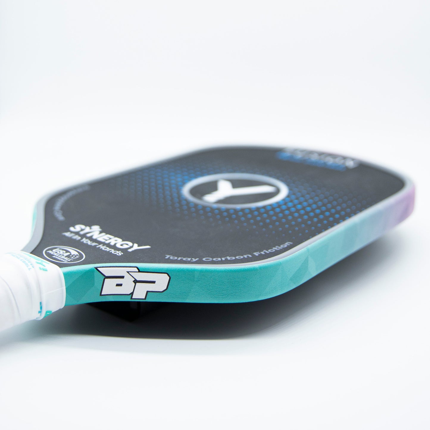 A close-up of a pickleball paddle with Bodhi Performance Premium Edge Tape in turquoise for professional edge guard protection, featuring a white grip and blue gradient face with SYNERGY and BP branding on a clean white background.