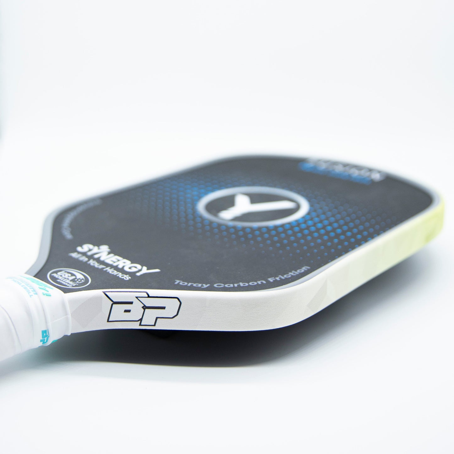 Close-up of a black and blue Bodhi Performance pickleball paddle with Premium Edge Tape for professional edge guard protection, carbon fiber texture, “SYNERGY” and “BP” branding, and a circular logo at the center, lying on a white surface.