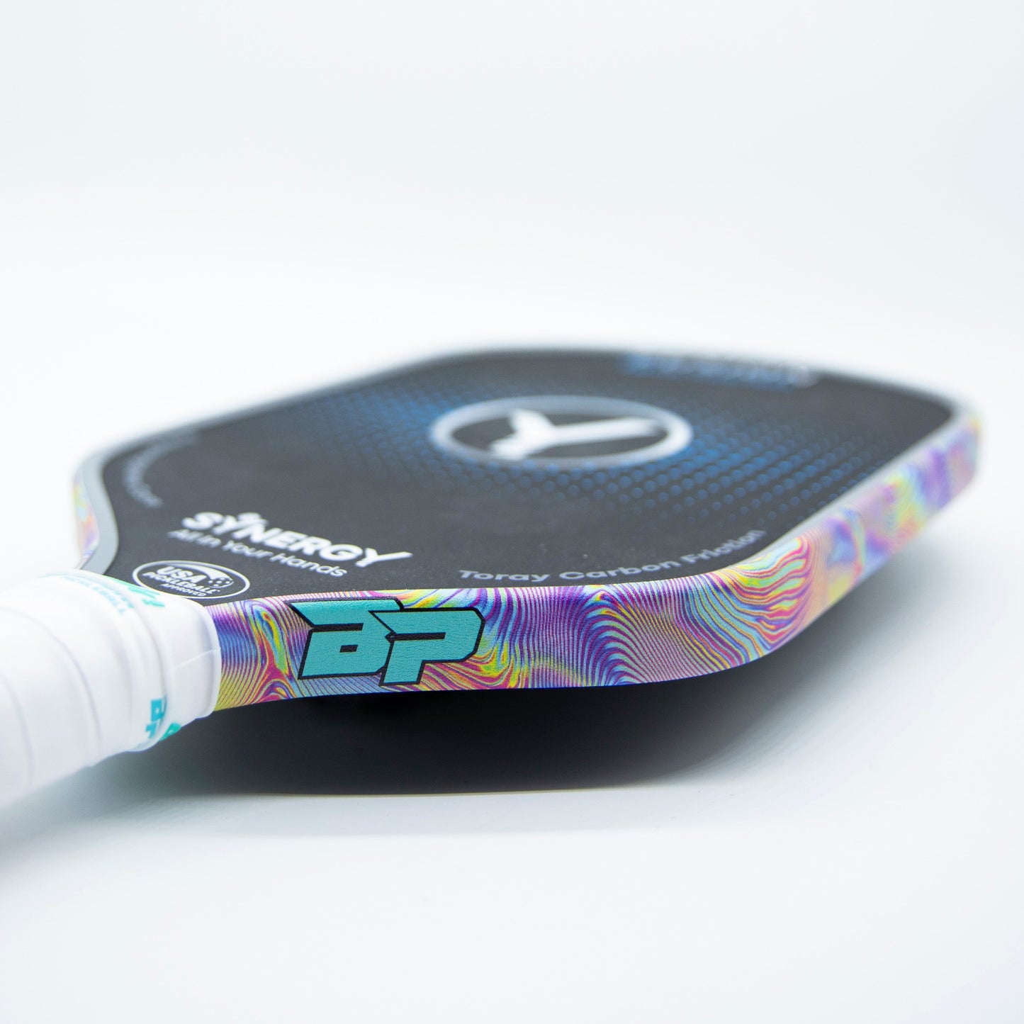 A close-up of a pickleball paddle on a white surface features a textured black face with a circular logo. Bodhi Performance - Premium Edge Tape provides colorful, wavy, professional edge guard protection, and the white handle has grip tape.