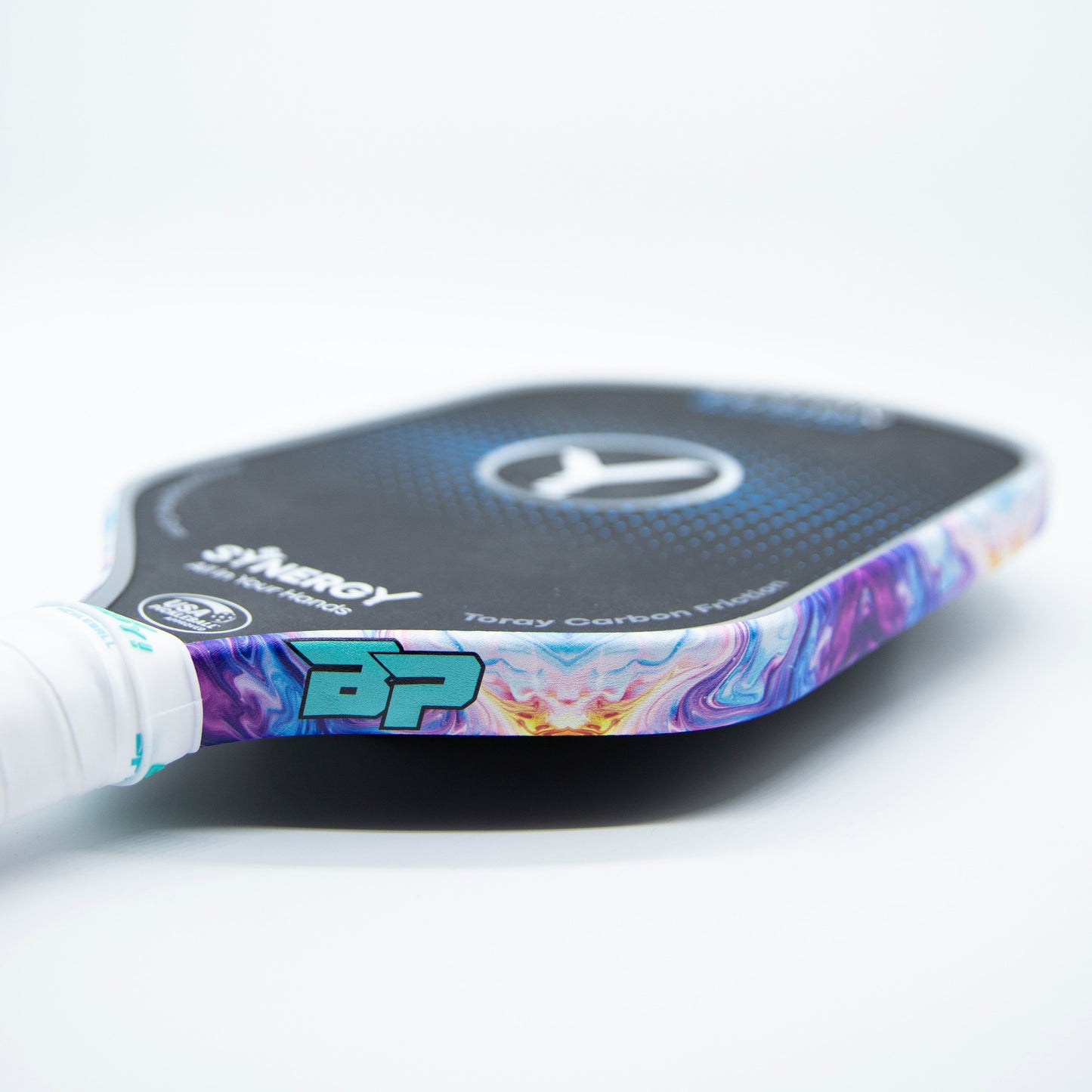 A close-up of a pickleball paddle with a white grip and Bodhi Performance Premium Edge Tape for professional edge guard protection, featuring a black carbon fiber face, the SYNERGY logo, and a turquoise Bodhi Performance (BP) logo near the handle.