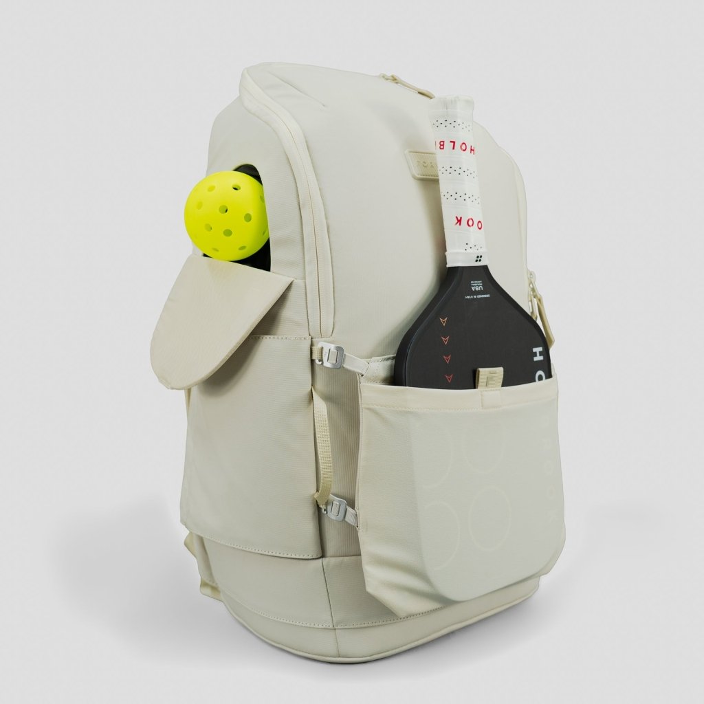 The FORWRD Court Caddy Front Paddle Sleeve in beige is shown with multiple pockets, a front sleeve holding a black paddle with a white handle in clear plastic, and a yellow perforated ball in the side pocket.