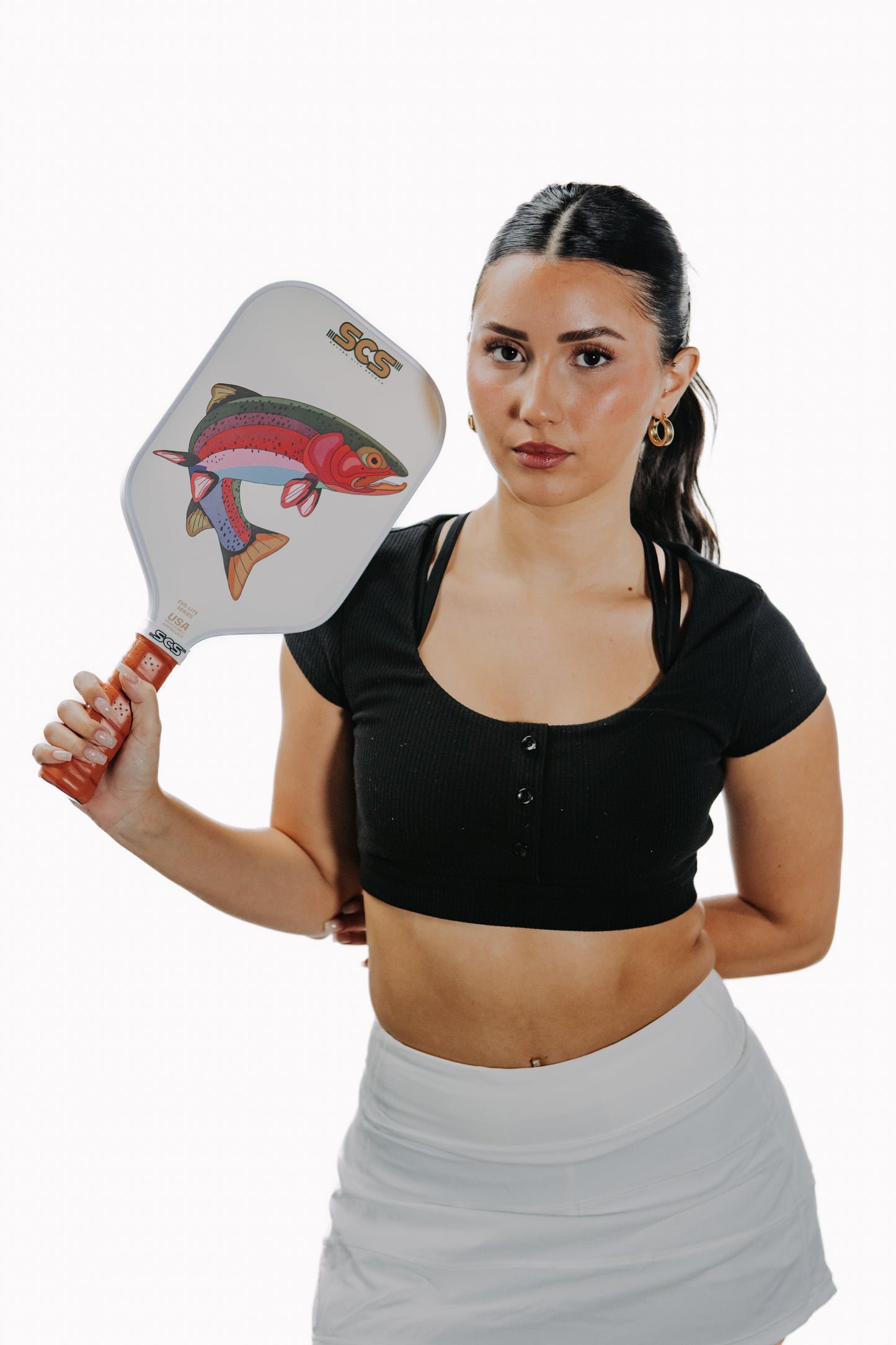 A woman with dark hair in a ponytail holds the Salted City Sports Bow Evo-Lite Series pickleball paddle, featuring a colorful fish design. She's in a black crop top and white skirt against a plain white background. USA Pickleball approved.
