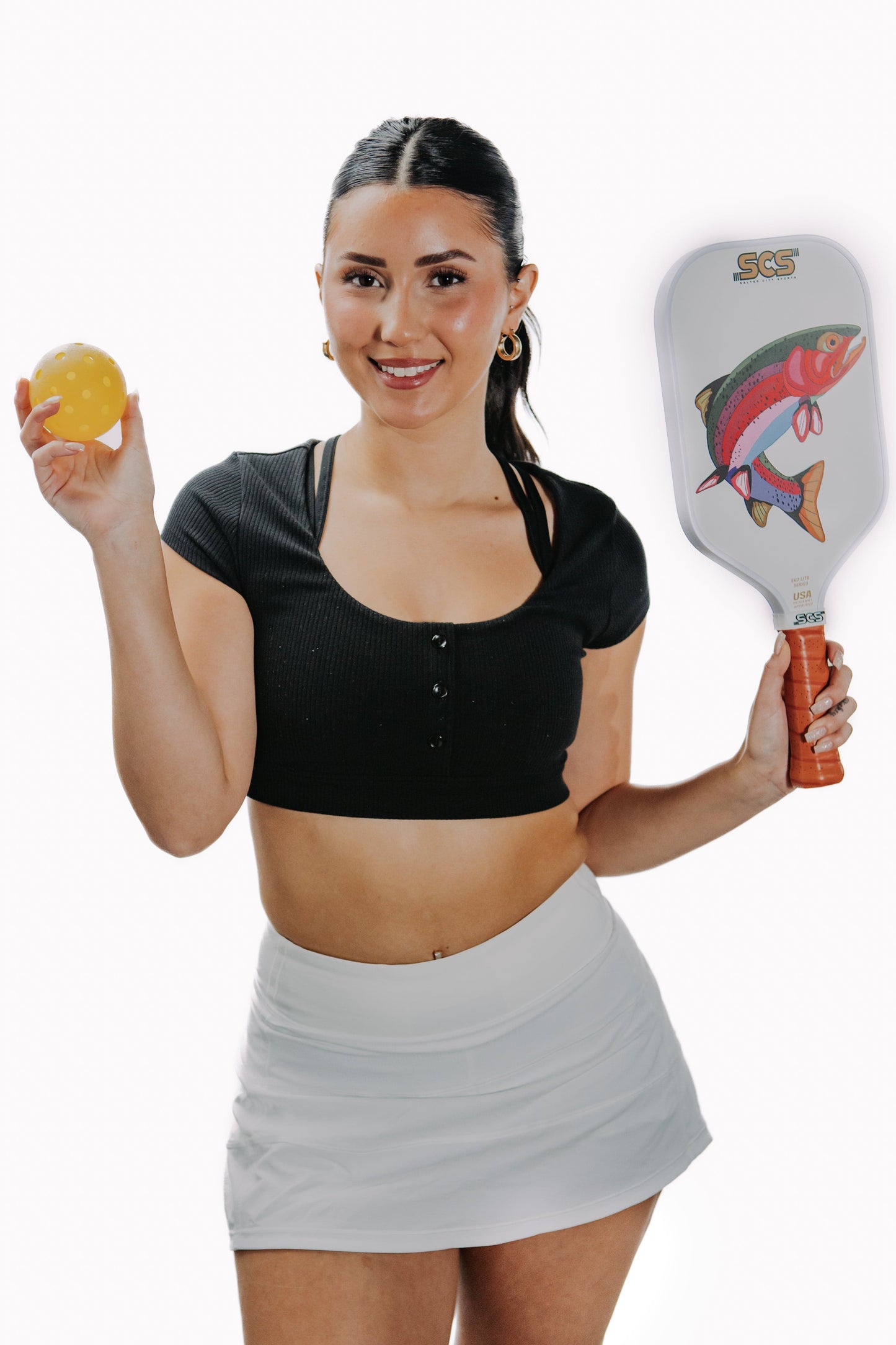 A smiling woman with dark hair in a ponytail wears a black crop top and white skirt, holding a yellow pickleball and a Salted City Sports Bow Evo-Lite Series paddle, USA Pickleball approved and decorated with a colorful fish graphic.