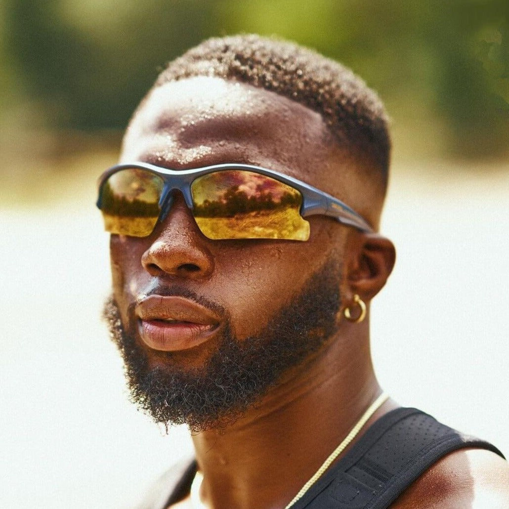 A man with a trimmed beard and faded haircut wears Epoch Eyewear's Brodie reflective sport sunglasses, a gold hoop earring, and a black tank top. Sunlight highlights his face against a blurred green outdoor background.