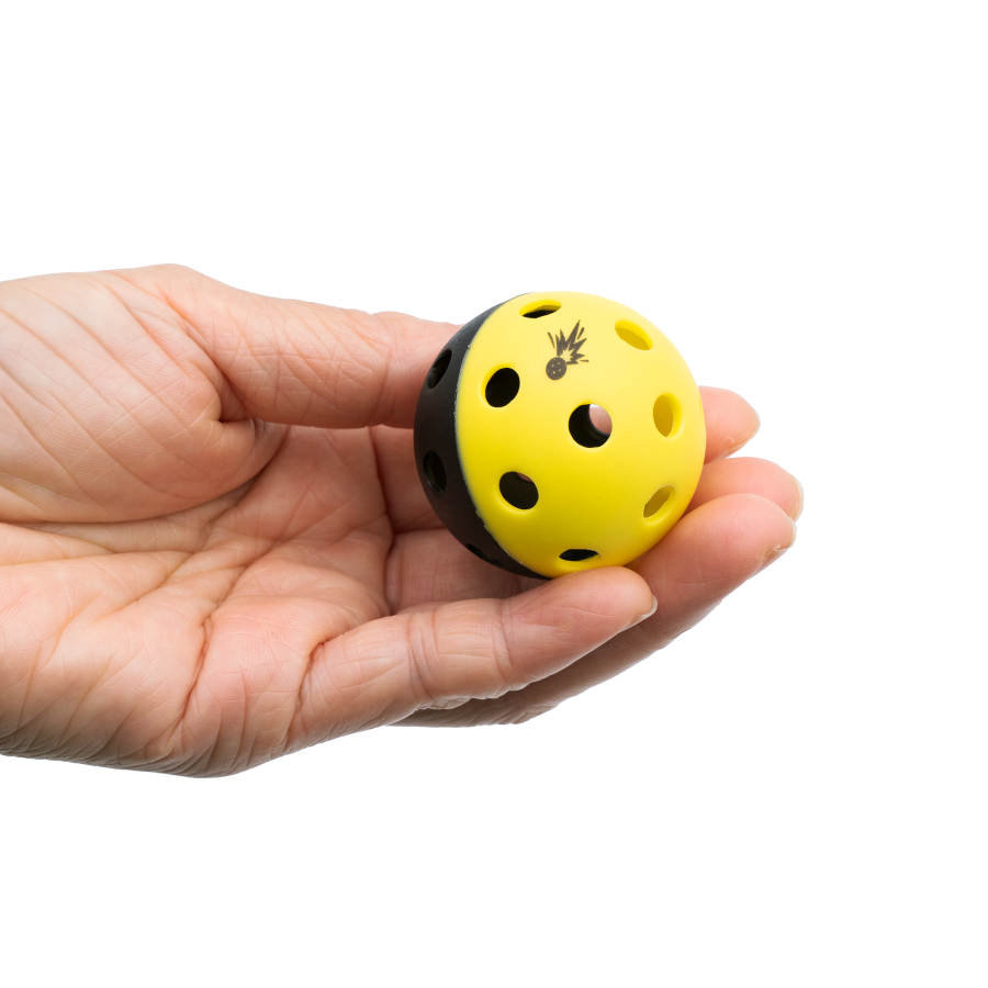 A left hand holds a Moment Pickleblasters 2-Color Mini Plastic Practice Ball by Moment Pickleball, half yellow and half black with holes, featuring a small black bee on the yellow side. The background is plain white.