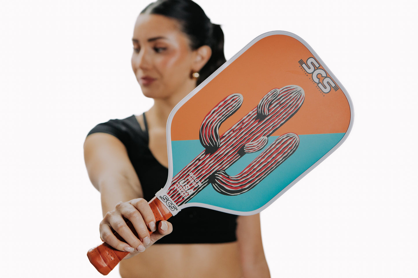 A woman in a black athletic top holds the Cacti | Evo-Lite Series paddle by Salted City Sports, showing its orange and teal design with cactus art and “SCS” lettering. The USA Pickleball approved paddle is held against a white background.