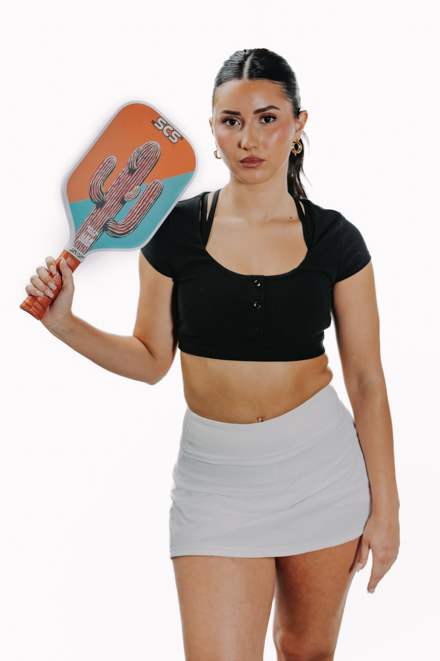 A woman with long dark hair, in a black crop top and light gray tennis skirt, holds the Salted City Sports Cacti | Evo-Lite Series pickleball paddle. She stands against a white background, gazing neutrally at the camera.