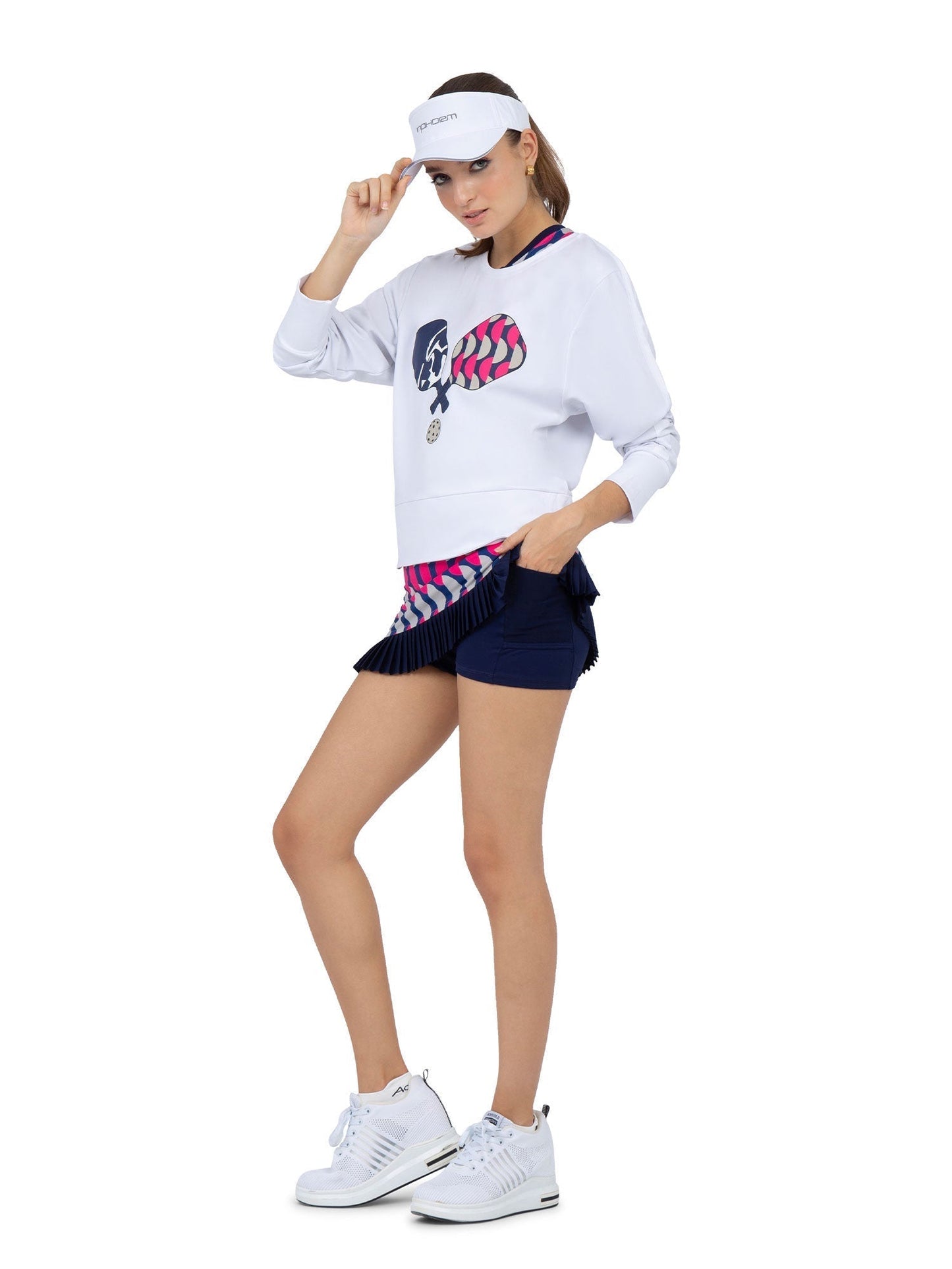 A woman poses in sporty style, wearing the inPhorm NYC Camila Performance Sweatshirt in Pickleball Print, a white visor, navy pleated skirt over shorts, and white sneakers. She tilts her visor with one hand against a plain white background.