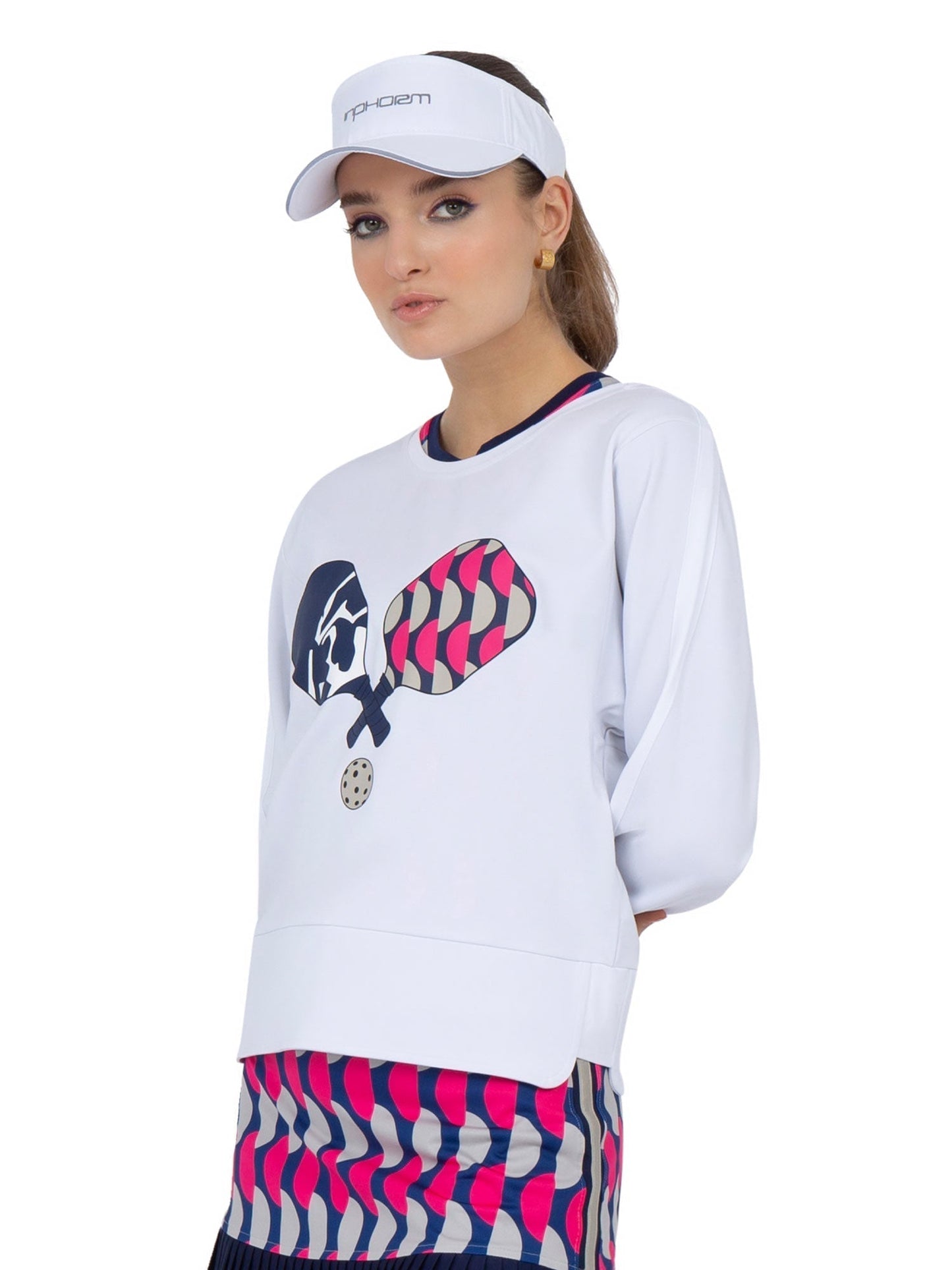 A woman in the inPhorm NYC Camila Performance Sweatshirt - Pickleball Print and a colorful geometric skirt stands slightly sideways, sporting a white sports visor. The sweatshirt features two paddles and a ball graphic.