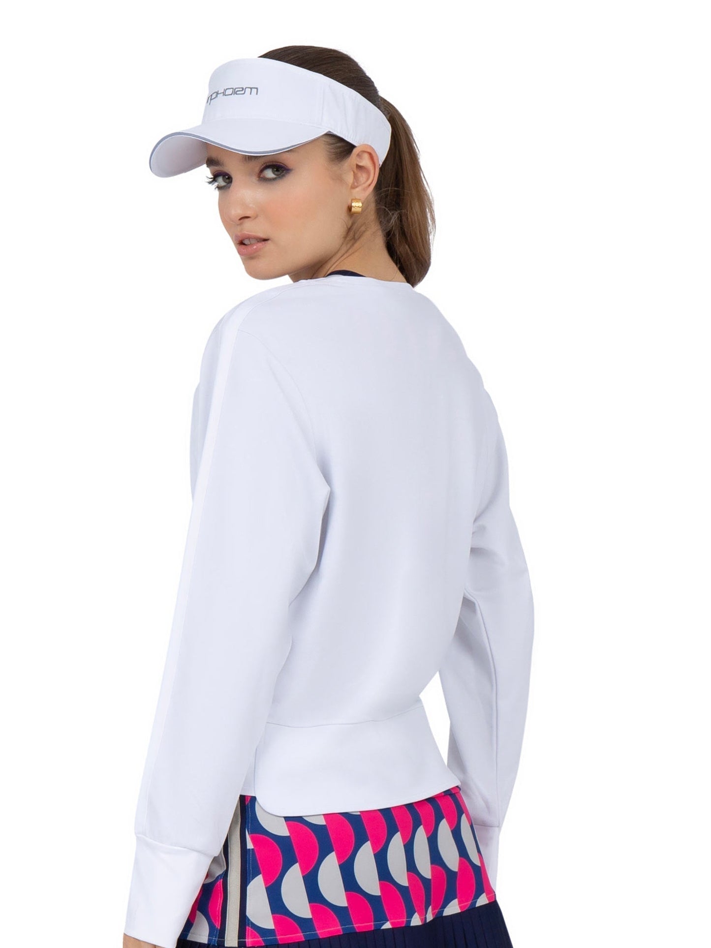 A woman in the inPhorm NYC Camila Performance Sweatshirt - Pickleball Print and visor looks over her shoulder. Her colorful geometric skirt pops against the plain white background, highlighting her sporty, stylish performance outfit.