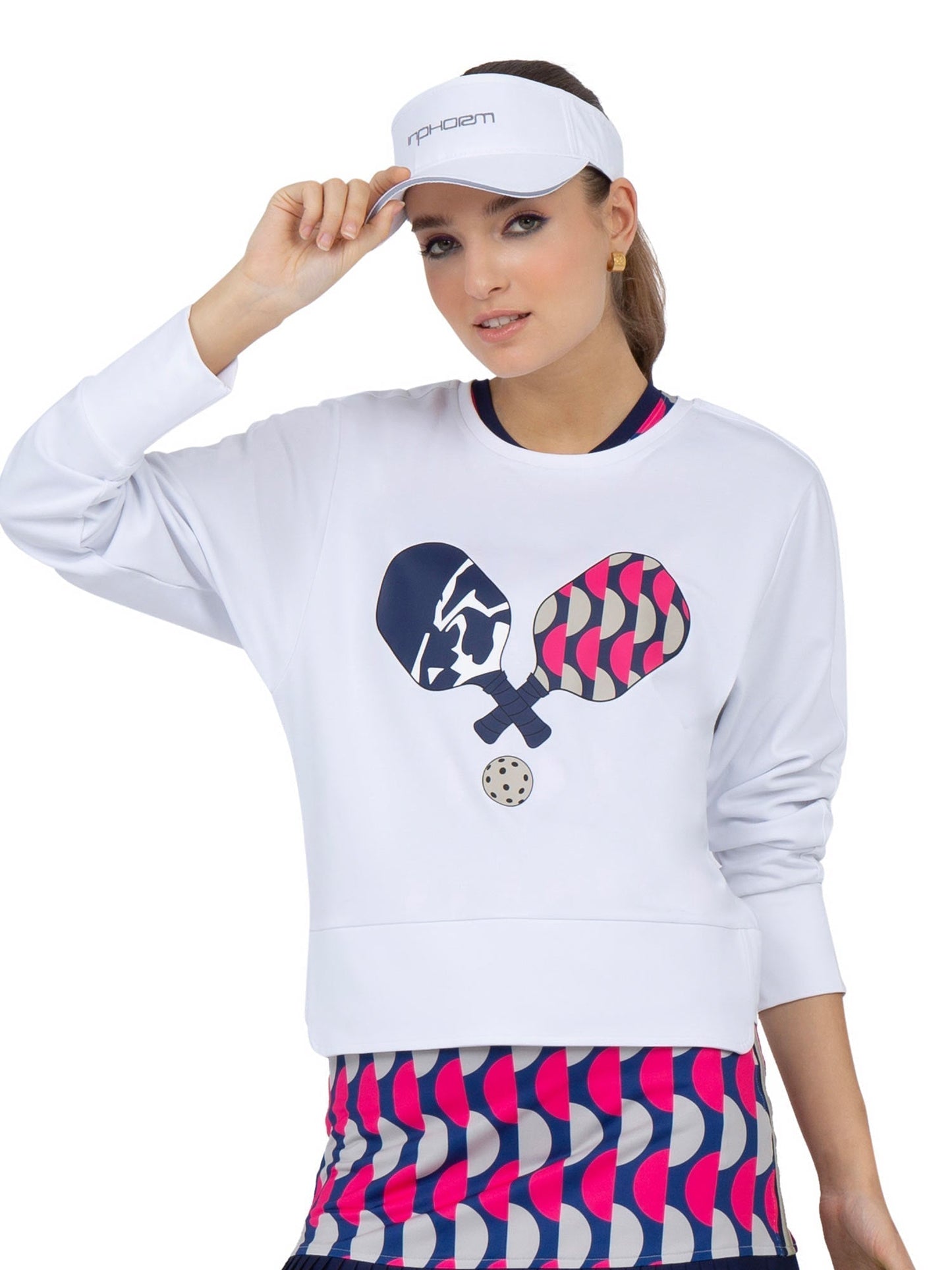 A woman wears the inPhorm NYC Camila Performance Sweatshirt - Pickleball Print, featuring two paddles and a ball, paired with a blue and pink wavy striped skirt. She adjusts her white visor and looks at the camera.