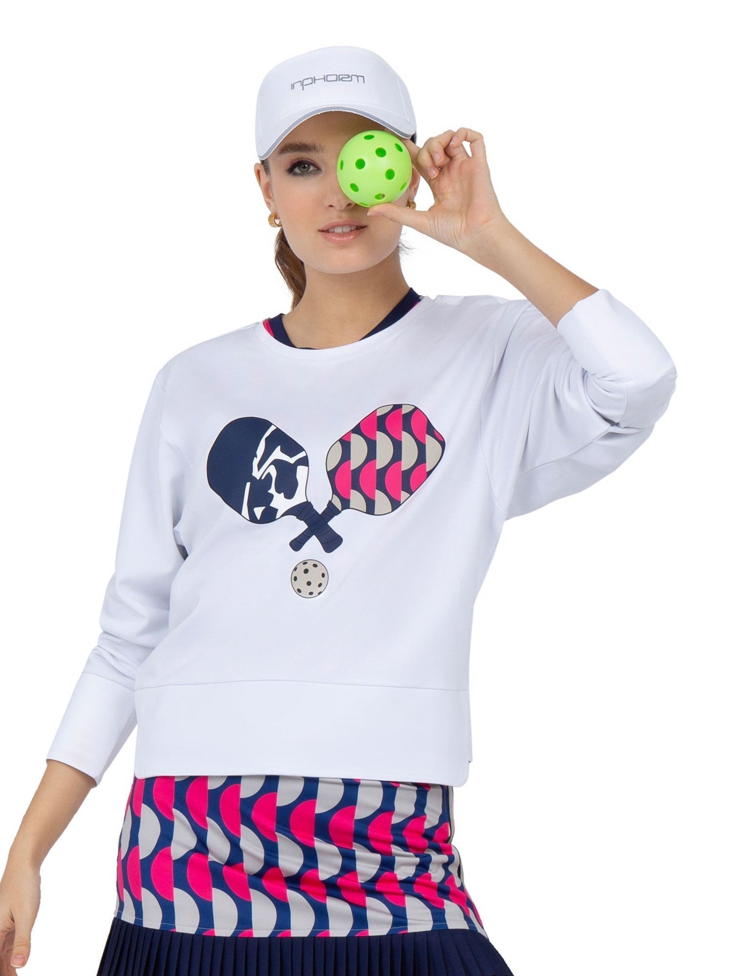 A woman in a white cap and the inPhorm NYC Camila Performance Sweatshirt – Pickleball Print holds a green pickleball over her eye. Her colorful skirt pops, while the UPF 50+ sweatshirt adds style and sun-safe, sporty cheer.