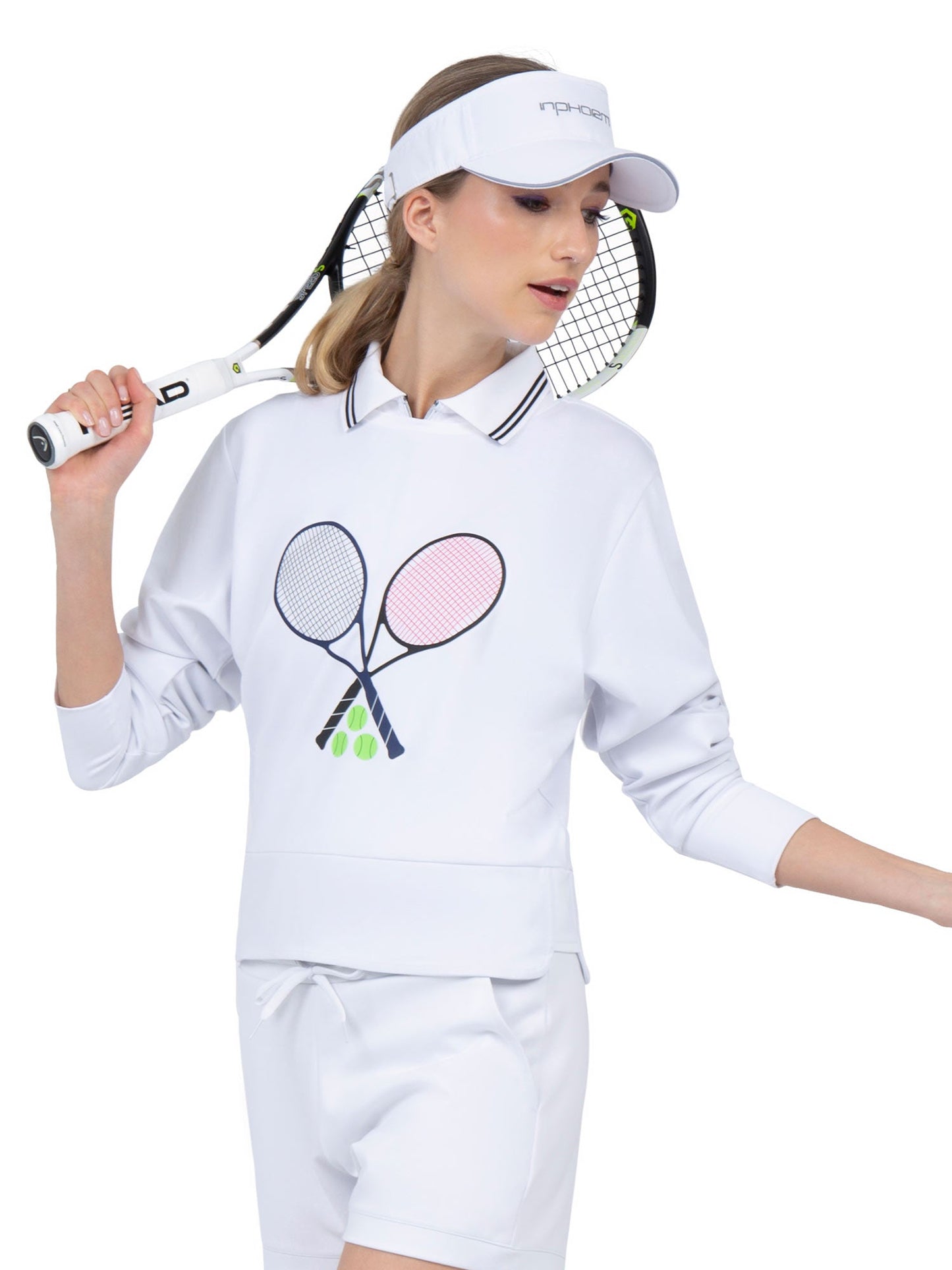 A woman in a white visor and the Camila Performance Sweatshirt - Tennis Print from inPhorm NYC, featuring UPF 50+ fabric, holds a racket over her shoulder, ready for tennis or pickleball with her hair in a low ponytail and white shorts.