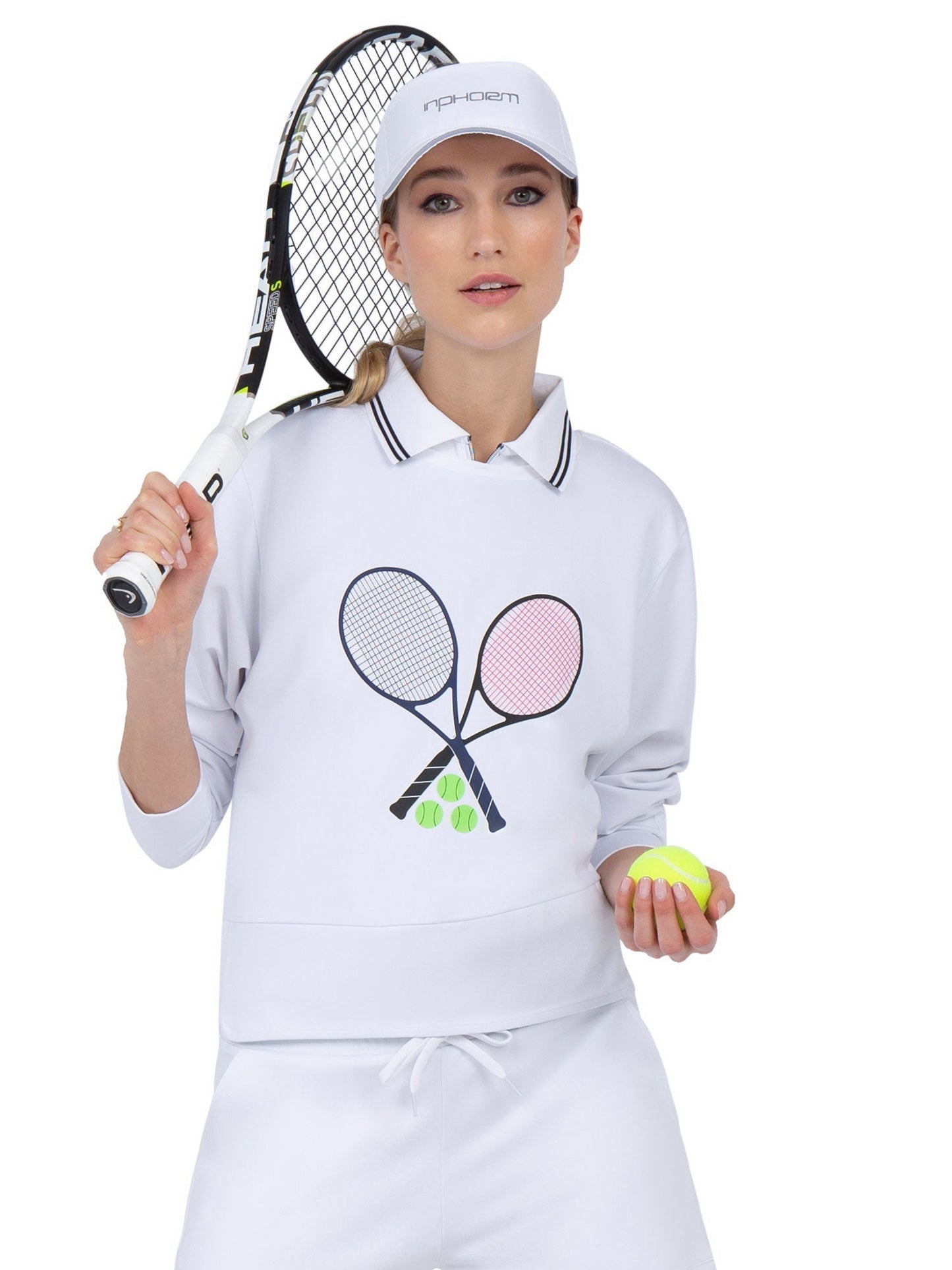 A woman in a white tennis outfit and cap holds a racket and a yellow ball. She wears the inPhorm NYC Camila Performance Sweatshirt – Tennis Print, featuring two crossed rackets and three green tennis balls on the front.