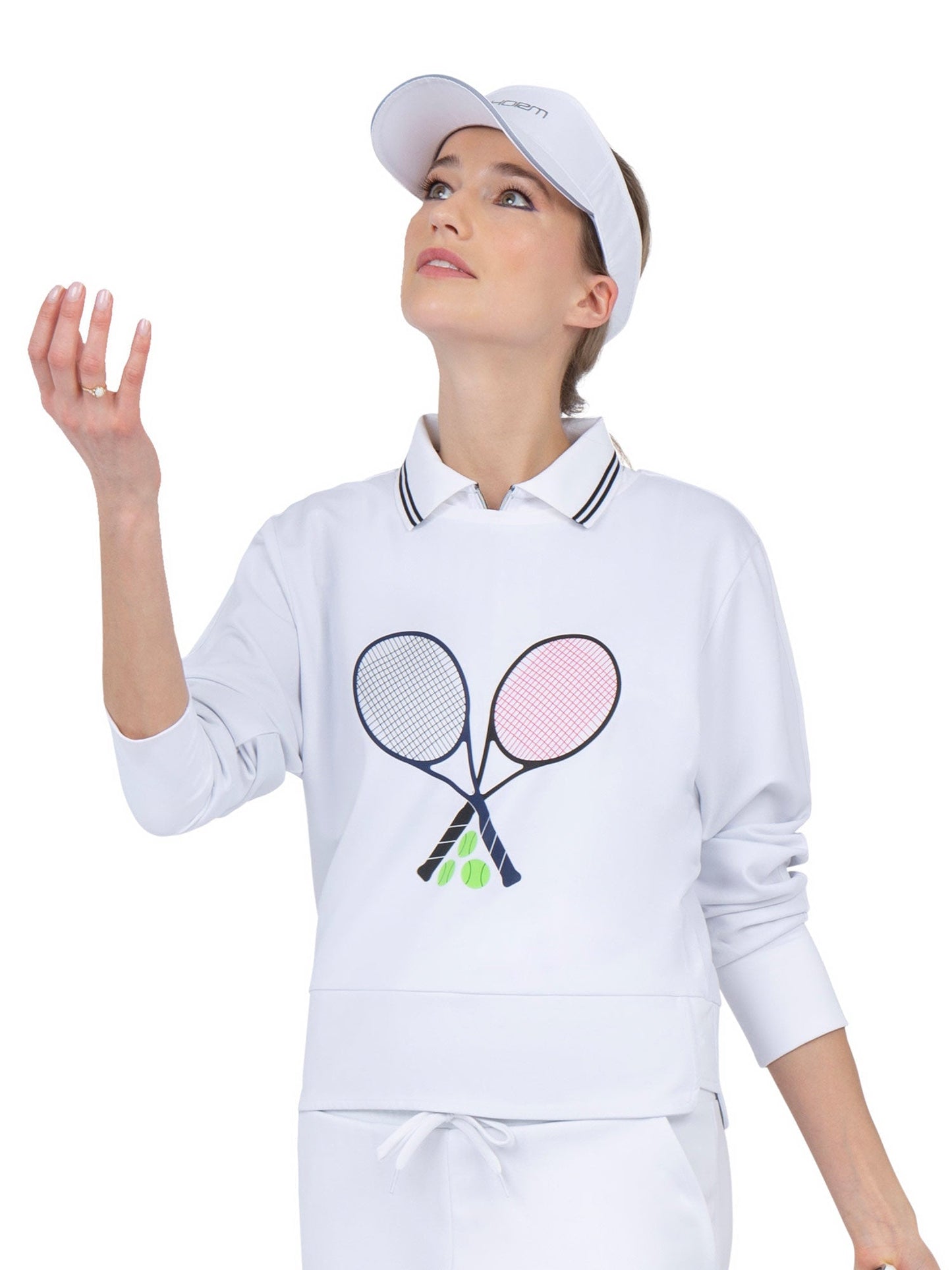 A woman in a white visor and the inPhorm NYC Camila Performance Sweatshirt - Tennis Print, featuring two crossed tennis rackets and tennis balls, looks upward while tossing a ball for a serve against a plain white background.