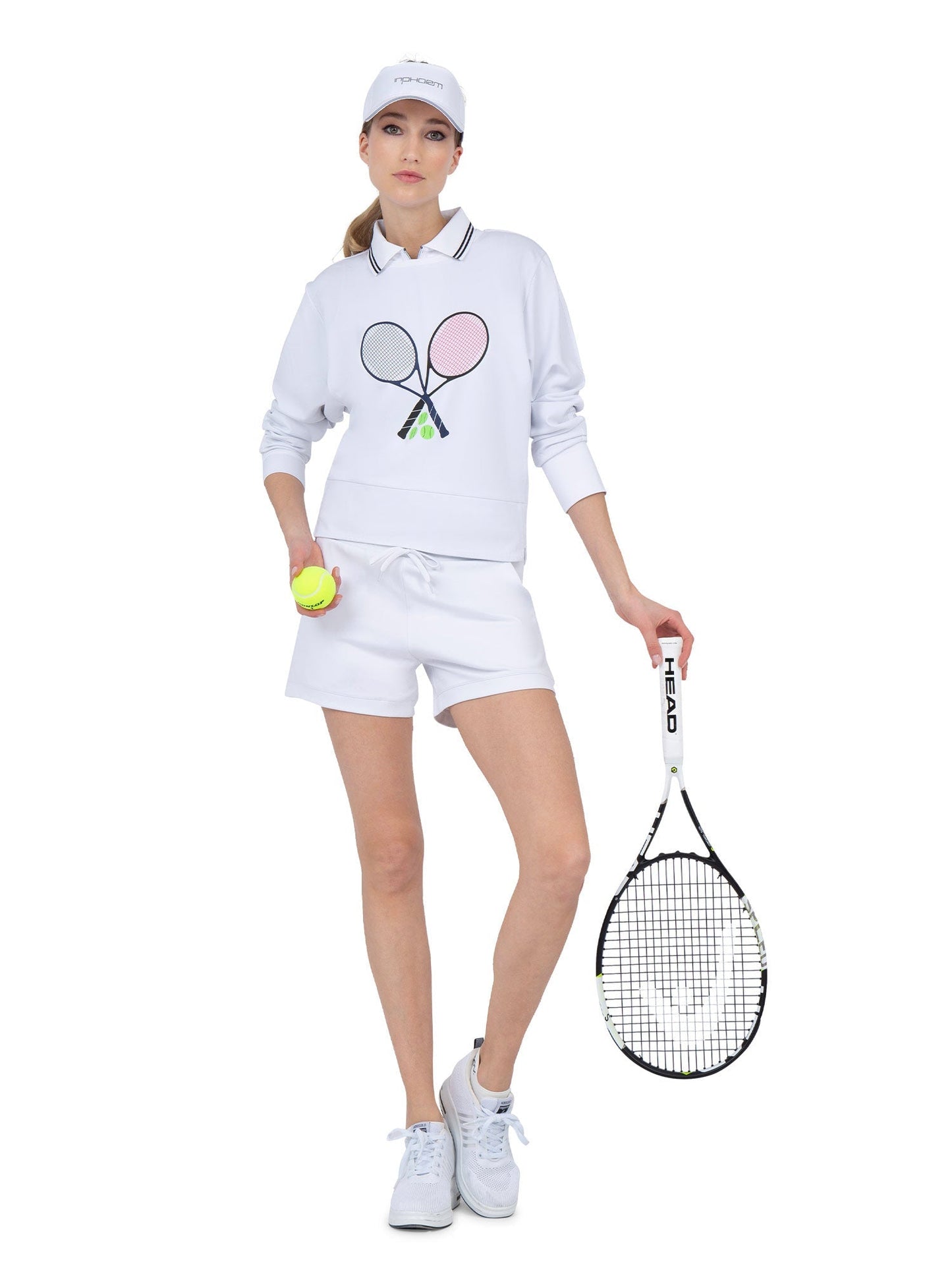 A woman in all-white tennis attire, including the inPhorm NYC Camila Performance Sweatshirt with a tennis print, holds a racket and yellow ball. She also wears a cap, shorts, and sneakers against a plain white background.