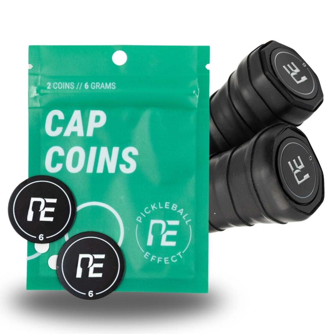 A green pouch labeled "Cap Coins" from Pickleball Effect Shop holds two black coins for paddle weight adjustment. Beside it are two paddle grips, each topped with a black PE-logo cap. The packaging shows the Pickleball Effect Shop brand.