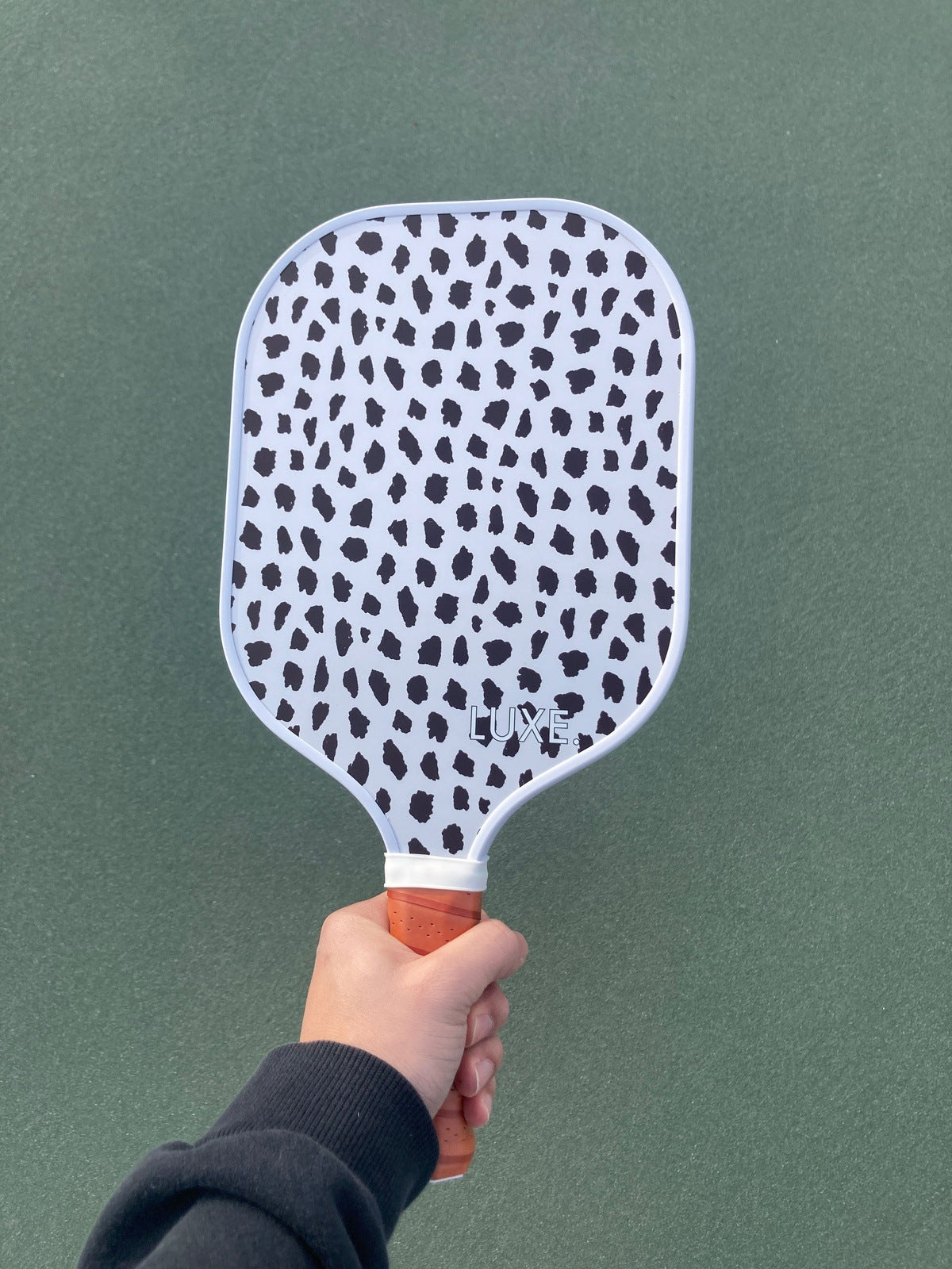 A person in a black sleeve holds up the LUXE Pickleball Cheetah paddle, featuring a white face with irregular black spots, orange grip, and brand name. The green pickleball court is visible in the background.