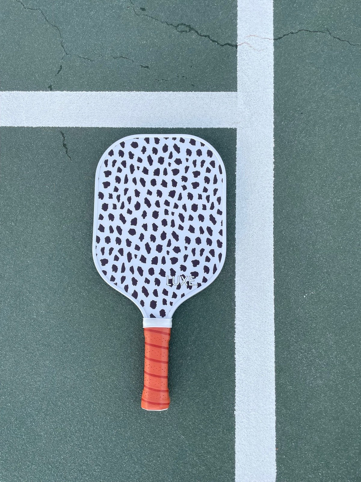 The LUXE Pickleball Cheetah paddle, featuring a cheetah print and orange grip band, rests at a right angle on the green court. Its striking pattern and design deliver impressive ball speed for every match.