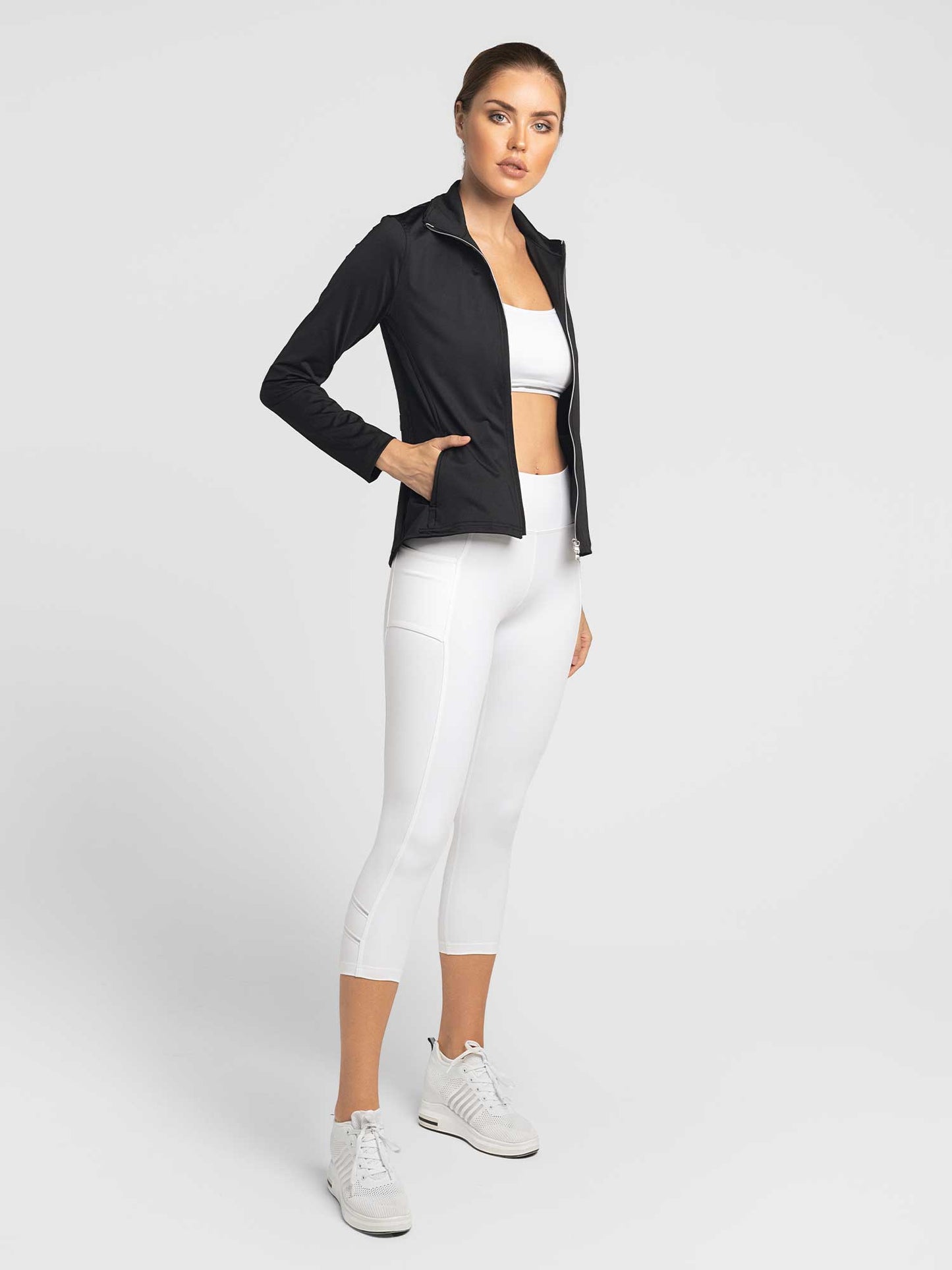 A woman with light skin and tied-back hair stands against a light gray background, wearing the inPhorm NYC Classic Blythe Jacket in Black over a white sports bra and high-waisted cropped leggings, paired with white sneakers and a neutral expression.