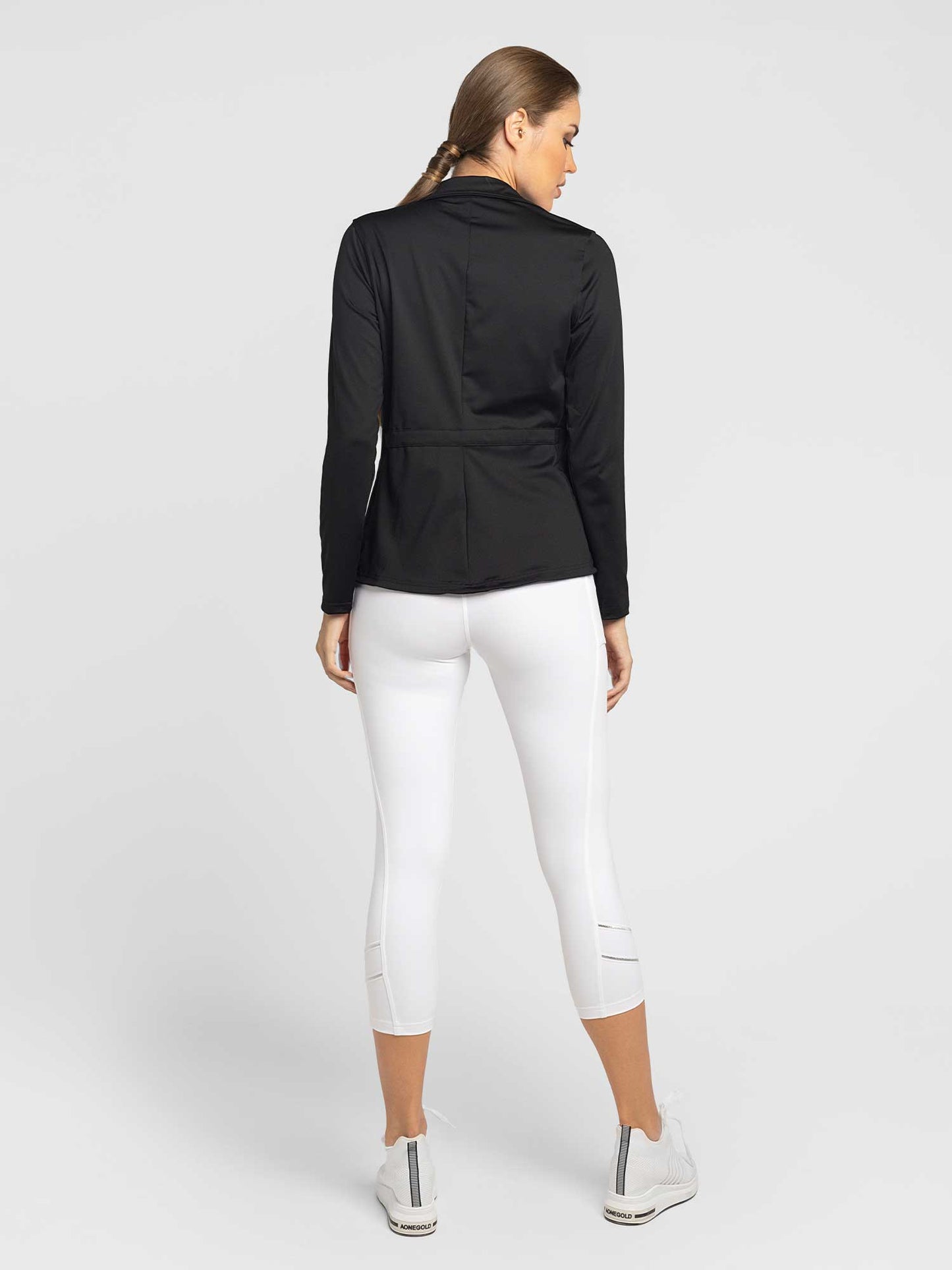 A woman with light brown hair in a low ponytail wears the inPhorm NYC Classic Blythe Jacket in black and white cropped leggings with white sneakers. The plain white background highlights her athletic, casual style.