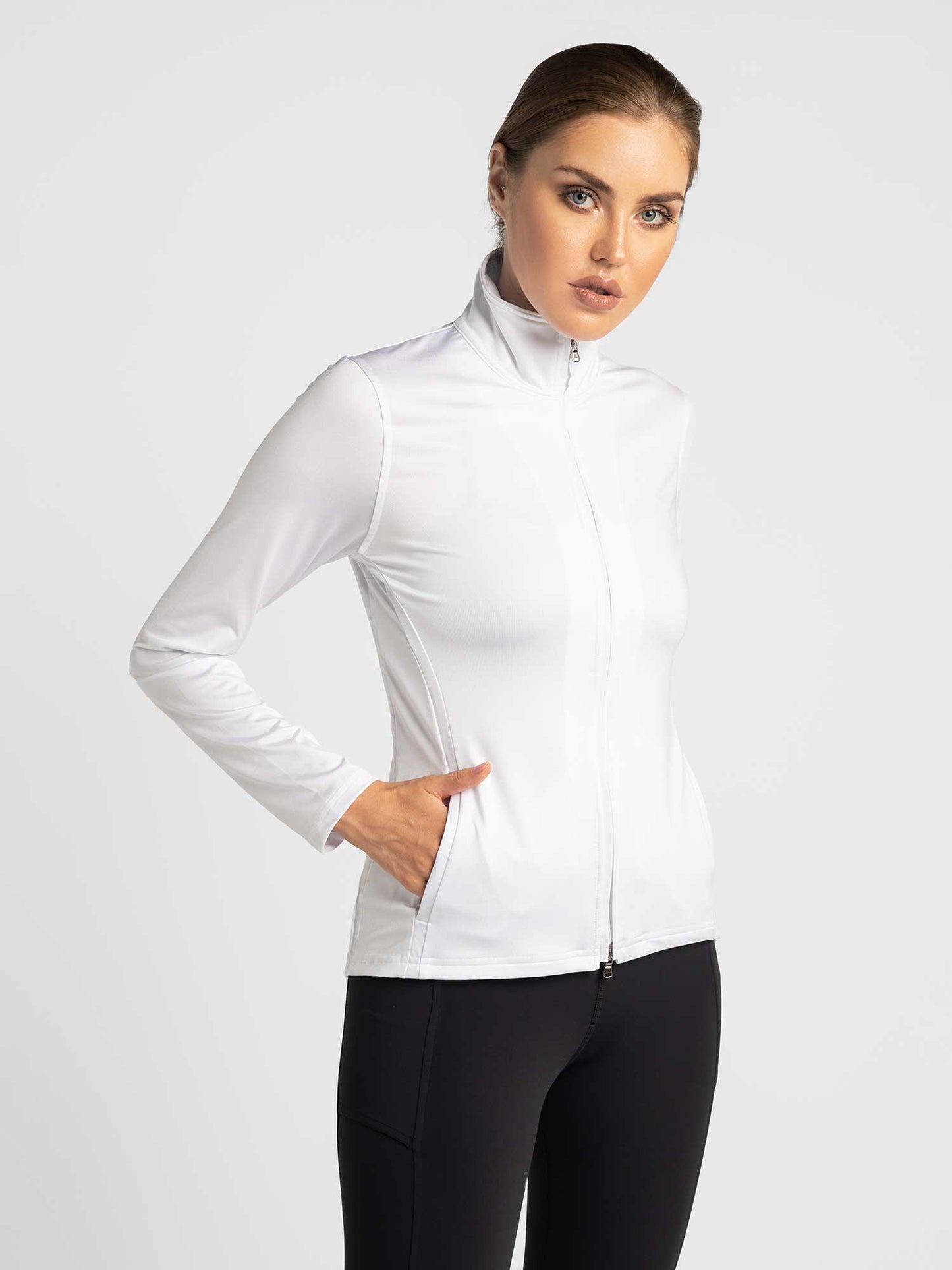 A woman with light skin and brown hair pulled back stands against a plain background, wearing the inPhorm NYC Classic Blythe Jacket in white and black leggings. She has one hand in her pocket and gazes slightly to the side with a neutral expression.
