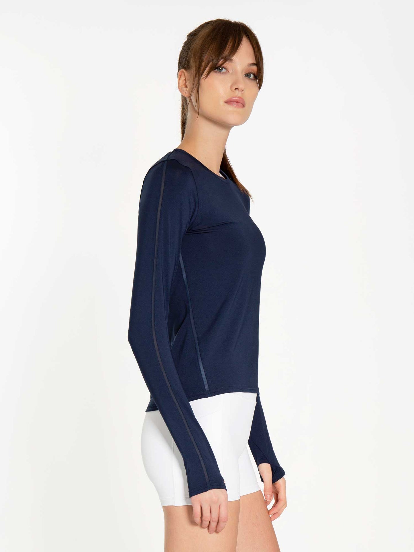 A woman with light skin and brown hair in a ponytail models the inPhorm NYC Classic Long Sleeve Crew - Midnight, a fitted moisture-wicking top, paired with white shorts. She stands in profile against a plain white background.