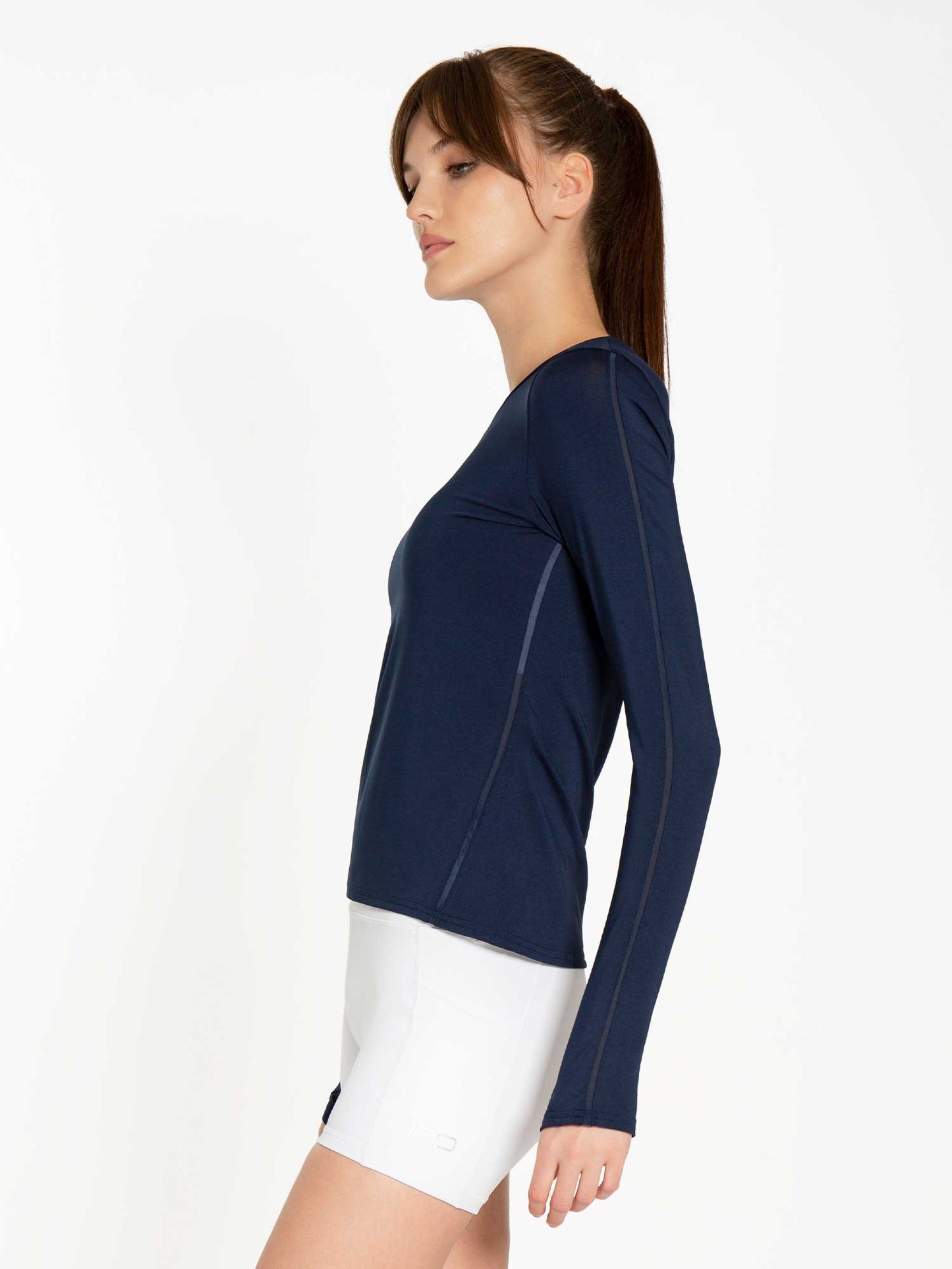 A woman with straight brown hair in a ponytail wears the inPhorm NYC Classic Long Sleeve Crew - Midnight (UPF 30-50+) and white athletic shorts. She stands in profile against a plain white background, looking calm.