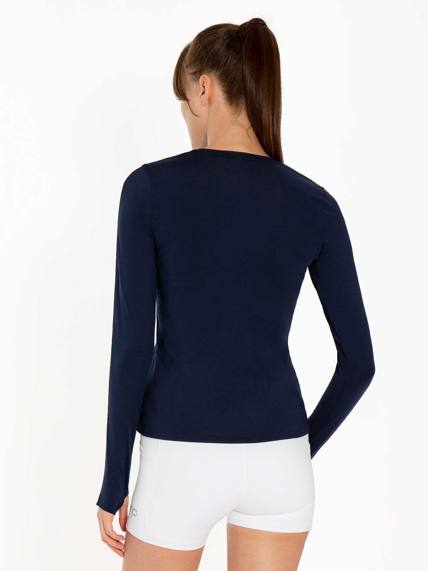 A woman with long brown hair in a ponytail faces away from the camera, wearing the inPhorm NYC Classic Long Sleeve Crew - Midnight and tight white shorts. A plain white backdrop and even lighting showcase her sporty, stylish outfit.