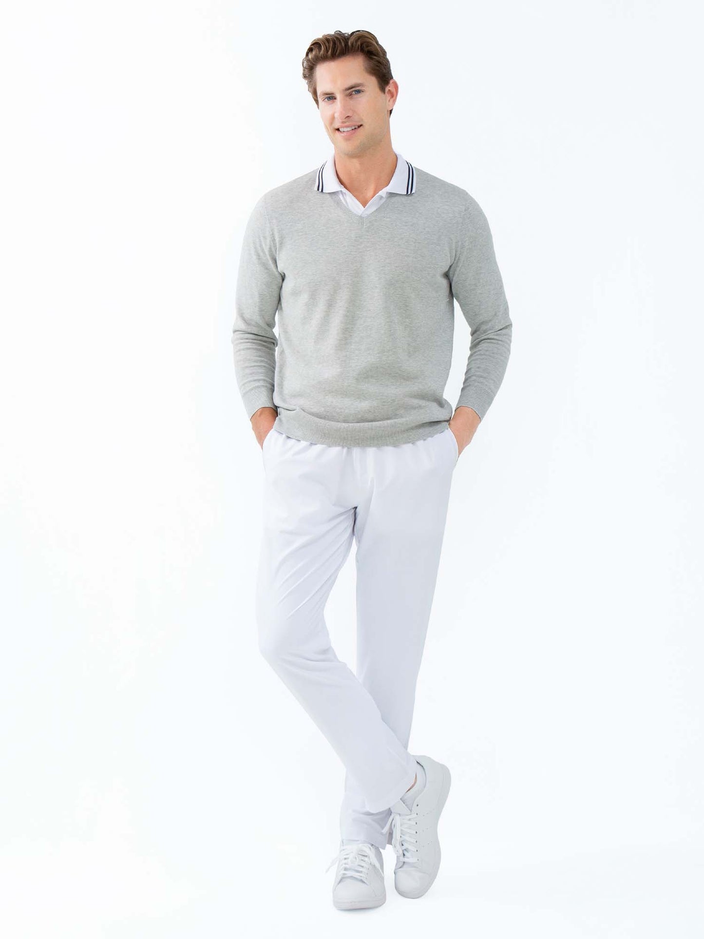 A man with short brown hair and a friendly expression stands against a white background wearing inPhorm NYC's Classic Performance Men's Tennis Pant in white, paired with a light gray V-neck sweater, white collared shirt, and white sneakers.