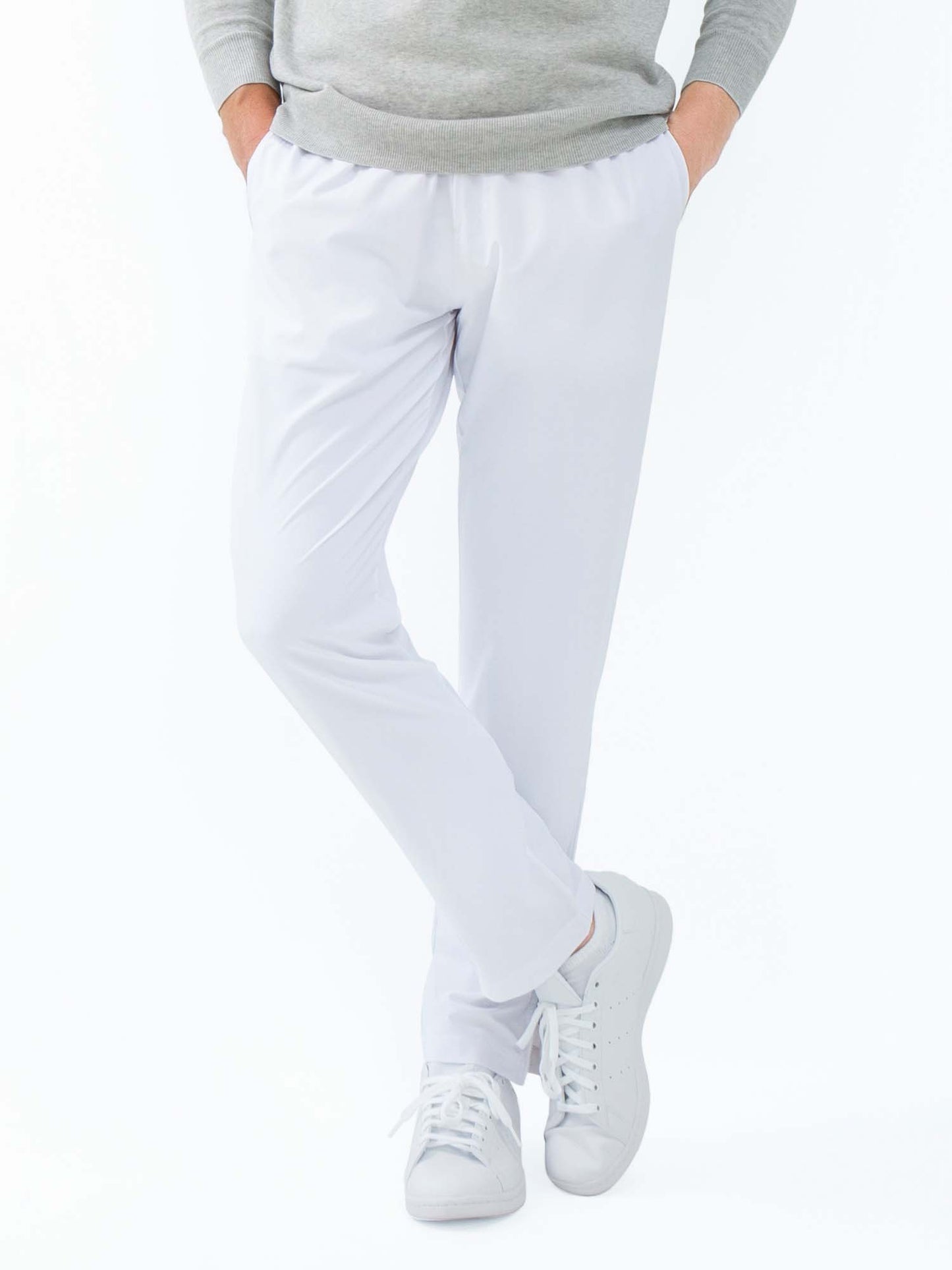 A person stands with legs crossed, hands in pockets, wearing inPhorm NYC's Classic Performance Men's Tennis Pant in white and a grey sweater, paired with white sneakers. The background is plain white. The person's head is not visible.