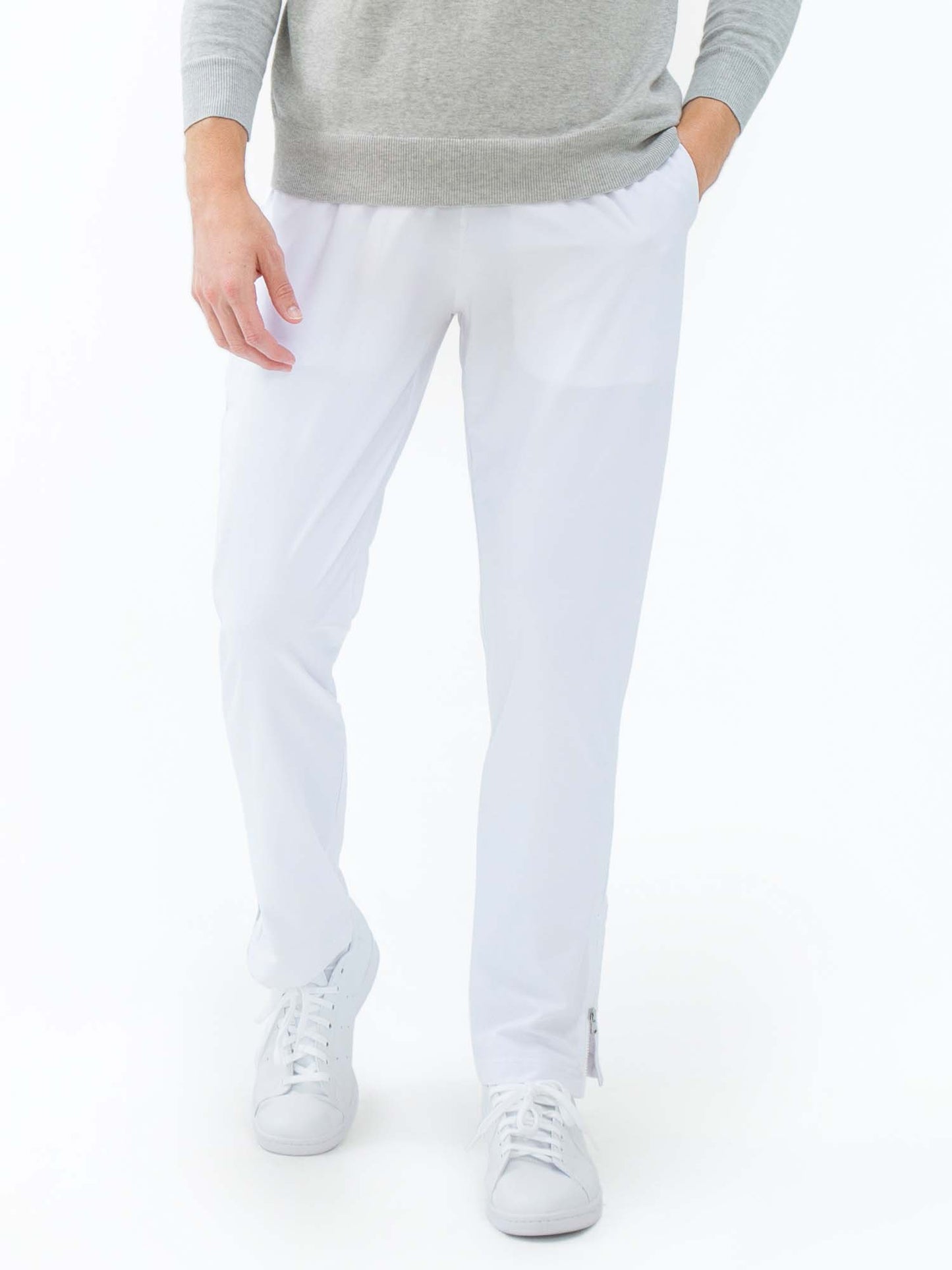 A person models the inPhorm NYC Classic Performance Men's Tennis Pant in white, featuring zippered ankles and performance stretch. Only the lower half is visible, with one hand at the side and white sneakers finishing the look against a plain background.