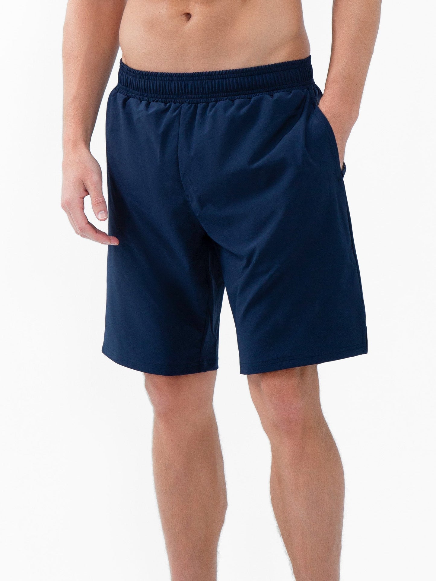 A man stands against a white background wearing the inPhorm NYC Classic Performance Men's Tennis Short in Midnight. The knee-length, relaxed fit shorts feature stretchy, quick-dry fabric and an elastic waistband; his right hand is partly in his pocket.