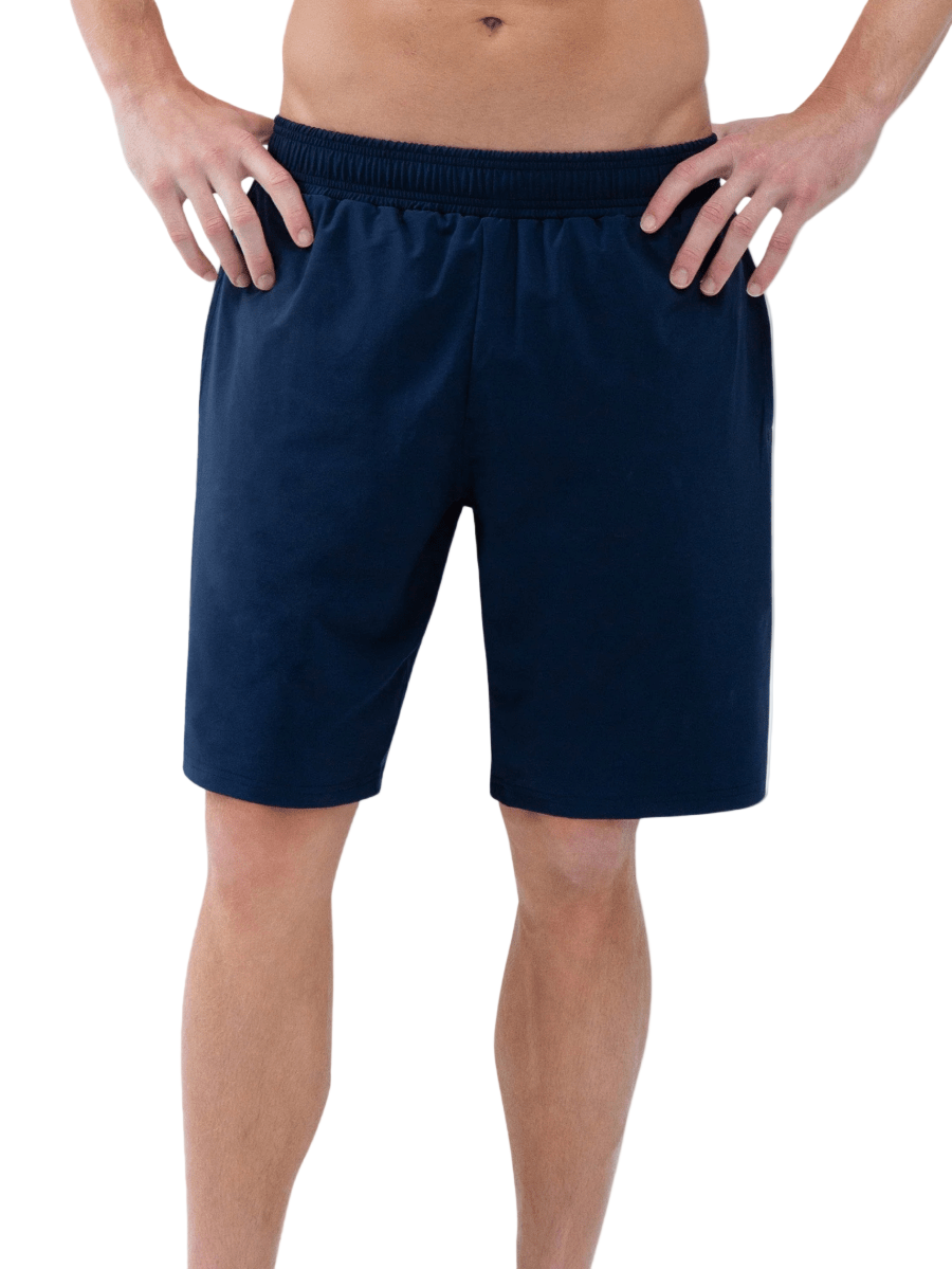 A person stands with hands on hips, wearing inPhorm NYC's Classic Performance Men's Tennis Short in Midnight. The photo, cropped from mid-torso to below the knees, features a smooth grey and white gradient background.