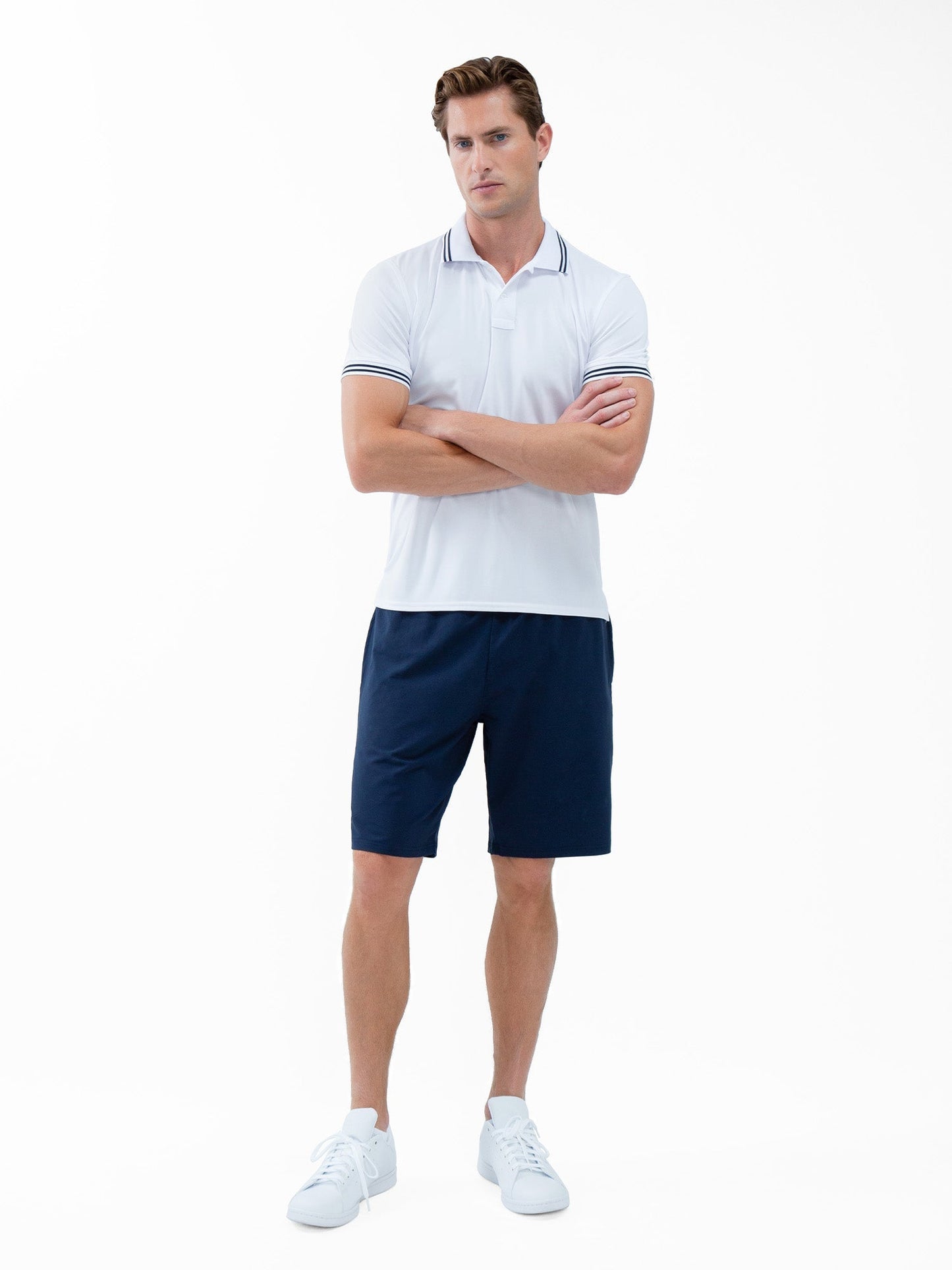 A man with short brown hair and a neutral expression stands with arms crossed, wearing inPhorm NYC’s Classic Performance Men's Tennis Short in Midnight, a white polo shirt with dark trim, and white sneakers. The background is plain white.