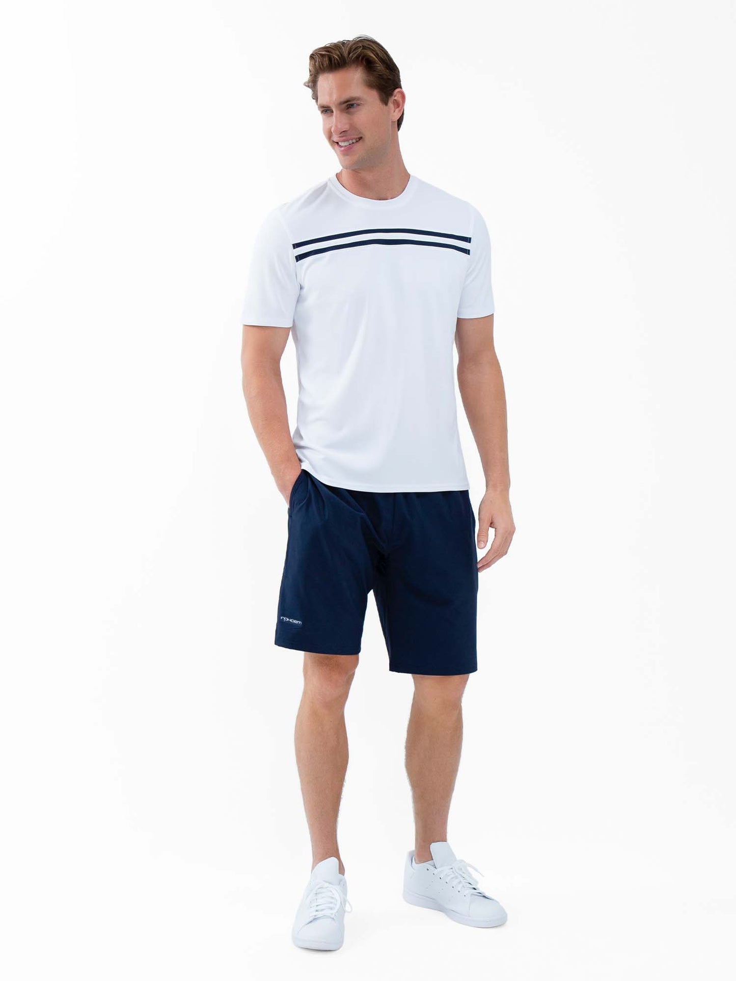 A man stands against a white background, smiling, wearing a white T-shirt with navy stripes, white sneakers, and the inPhorm NYC Classic Performance Men's Tennis Short in Midnight. One hand is in his pocket; the other rests by his side.