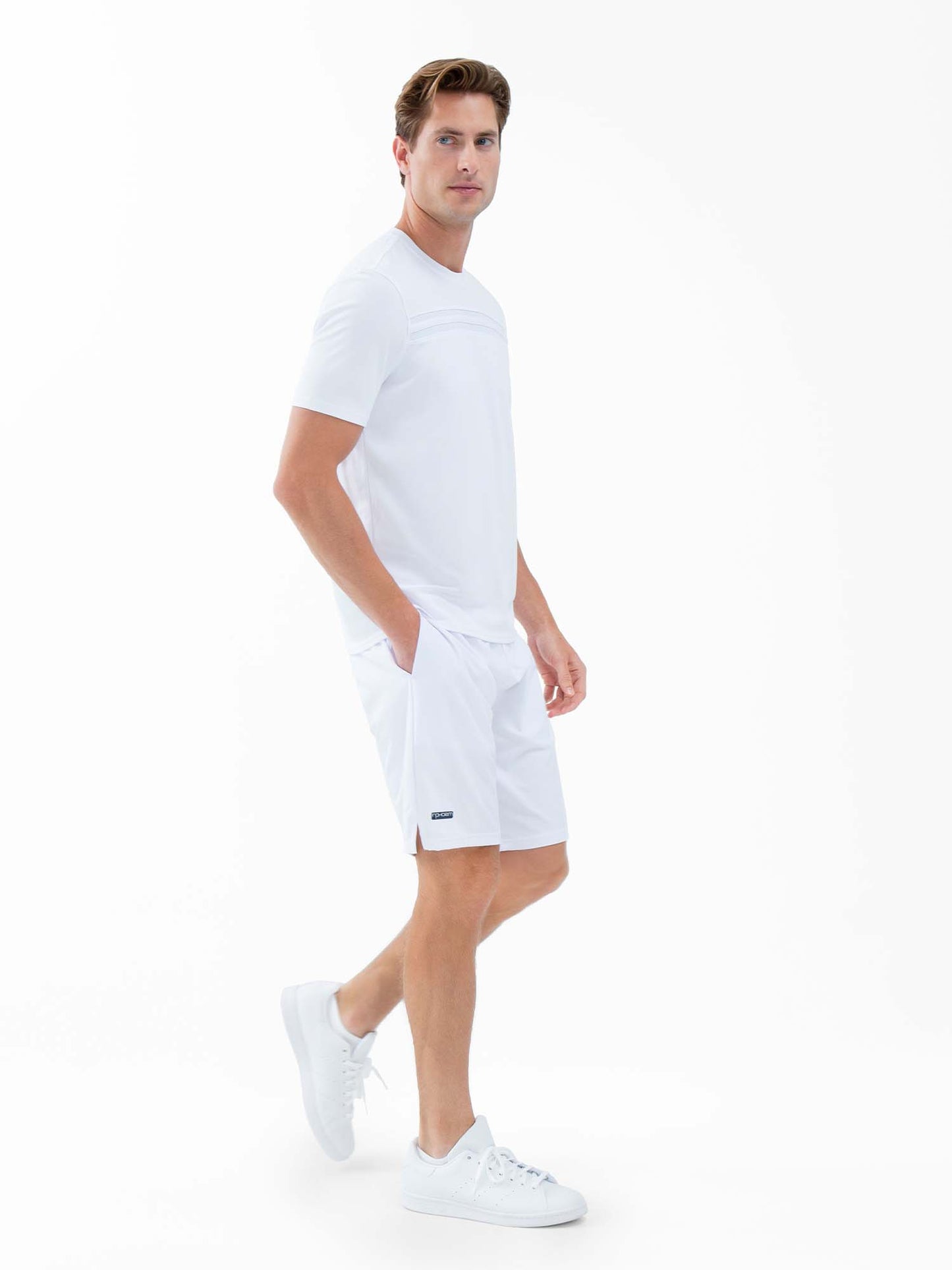 A man with light skin and short brown hair stands against a plain white background, wearing inPhorm NYC's Classic Performance Men's Tennis Short in white, featuring a built-in liner and quick-dry fabric. His hands are in his pockets as he gazes slightly right.