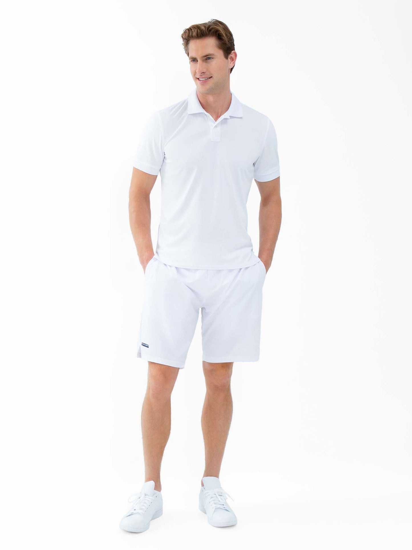 A man with neatly styled light brown hair stands against a white background, wearing inPhorm NYC's Classic Performance Men's Tennis Short in white with a built-in liner and quick-dry fabric. His hands are in his pockets and he smiles slightly.
