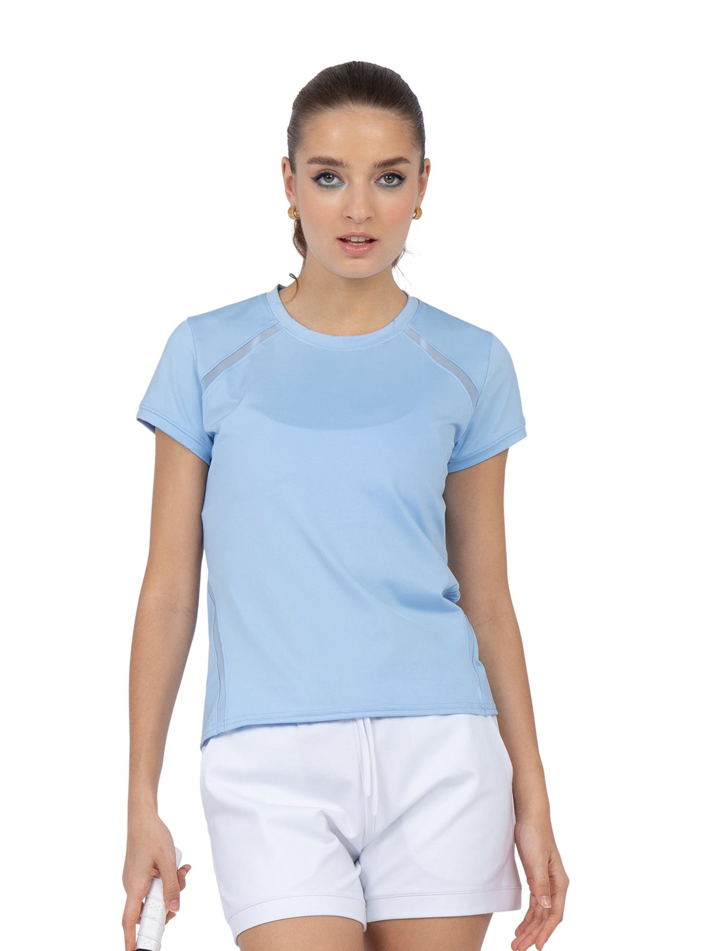 A woman with light skin and brown hair wears the inPhorm NYC Classic Short Sleeve Crew Neck in Bluebell and white shorts. She stands against a white background, holding a small white item, looking at the camera with a neutral expression.