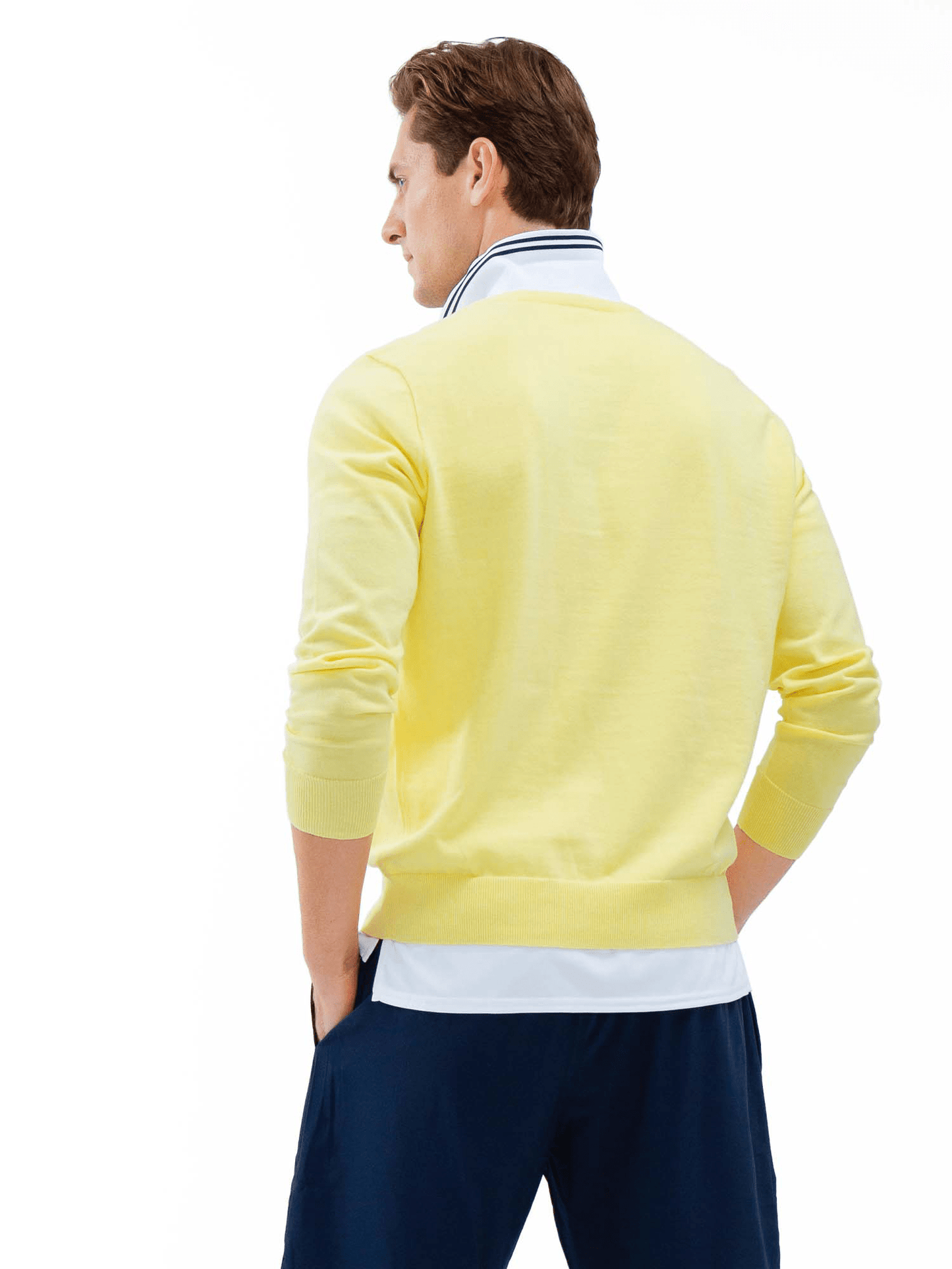 A man with neatly styled short brown hair stands facing away, hands in pockets. He wears the inPhorm NYC Classic V Neck Sweater in Lemonade over a white collared shirt with dark trim and navy blue pants against a plain white background.