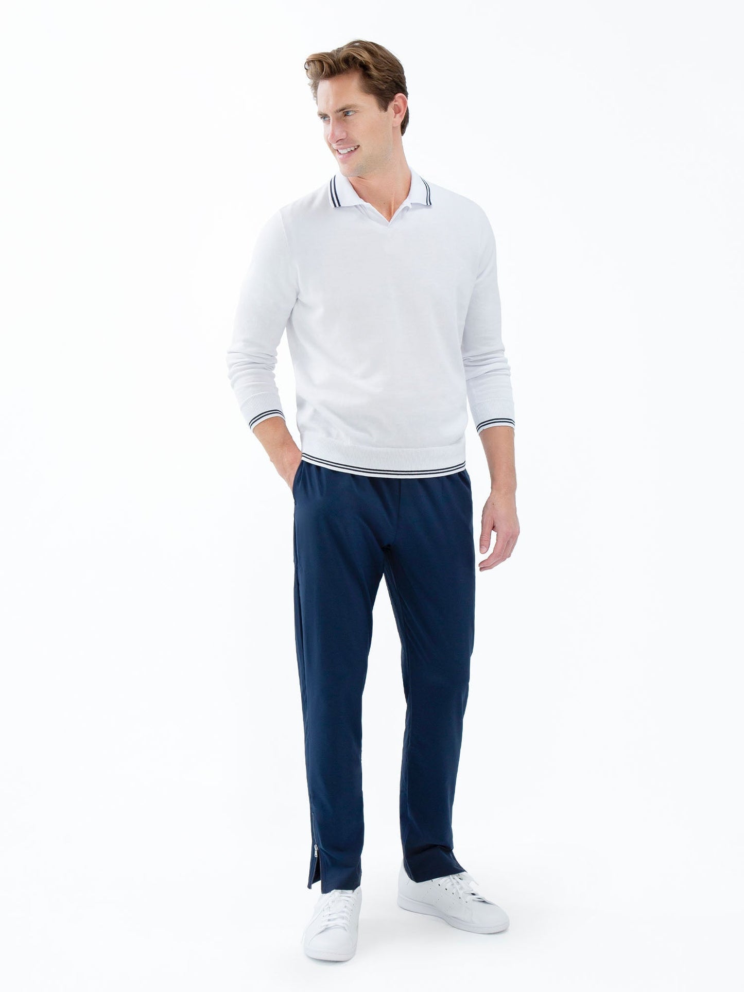 A man stands against a white background, smiling, wearing the inPhorm NYC Classic V Neck Sweater in White/Midnight. He pairs it with navy pants and white sneakers, looking slightly to the side with his arms relaxed by his sides.