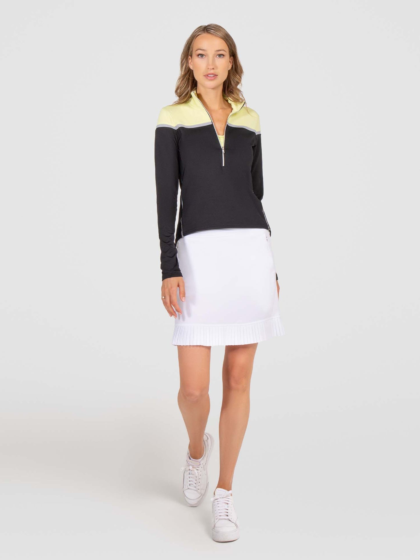 A woman with loose, wavy light brown hair stands against a white background, wearing inPhorm NYC’s Cleo 17" Golf Skirt in white—featuring built-in shorts and moisture-wicking fabric—with a black and light yellow long-sleeve zip-up top.