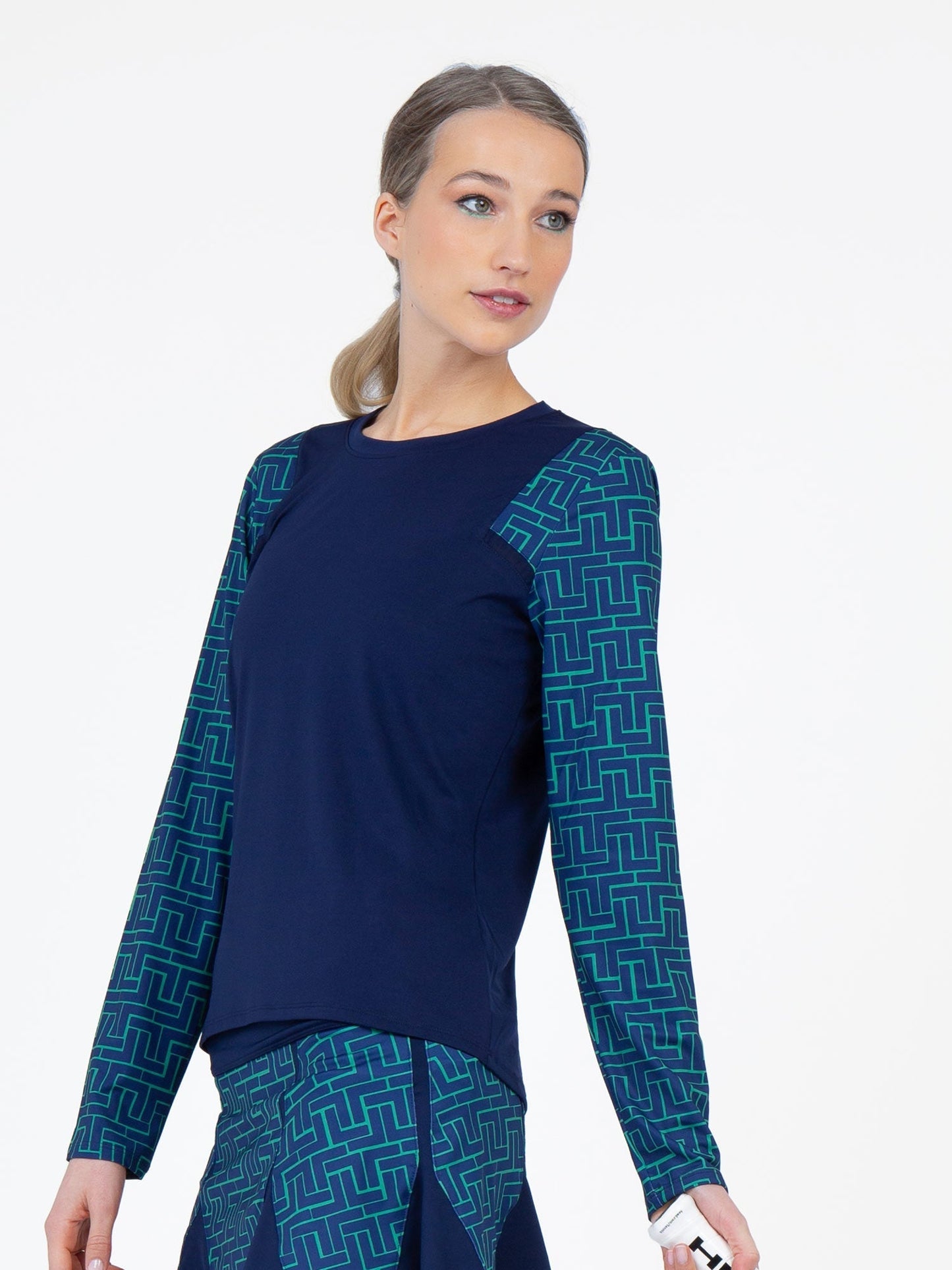 A woman with blond hair in a low ponytail models the inPhorm NYC Colorblock Active Sofia Long Sleeve Crew Neck in Ink/Ink Tessel, showcasing navy with green and blue geometric sleeve patterns and a matching skirt, set against a plain white background.
